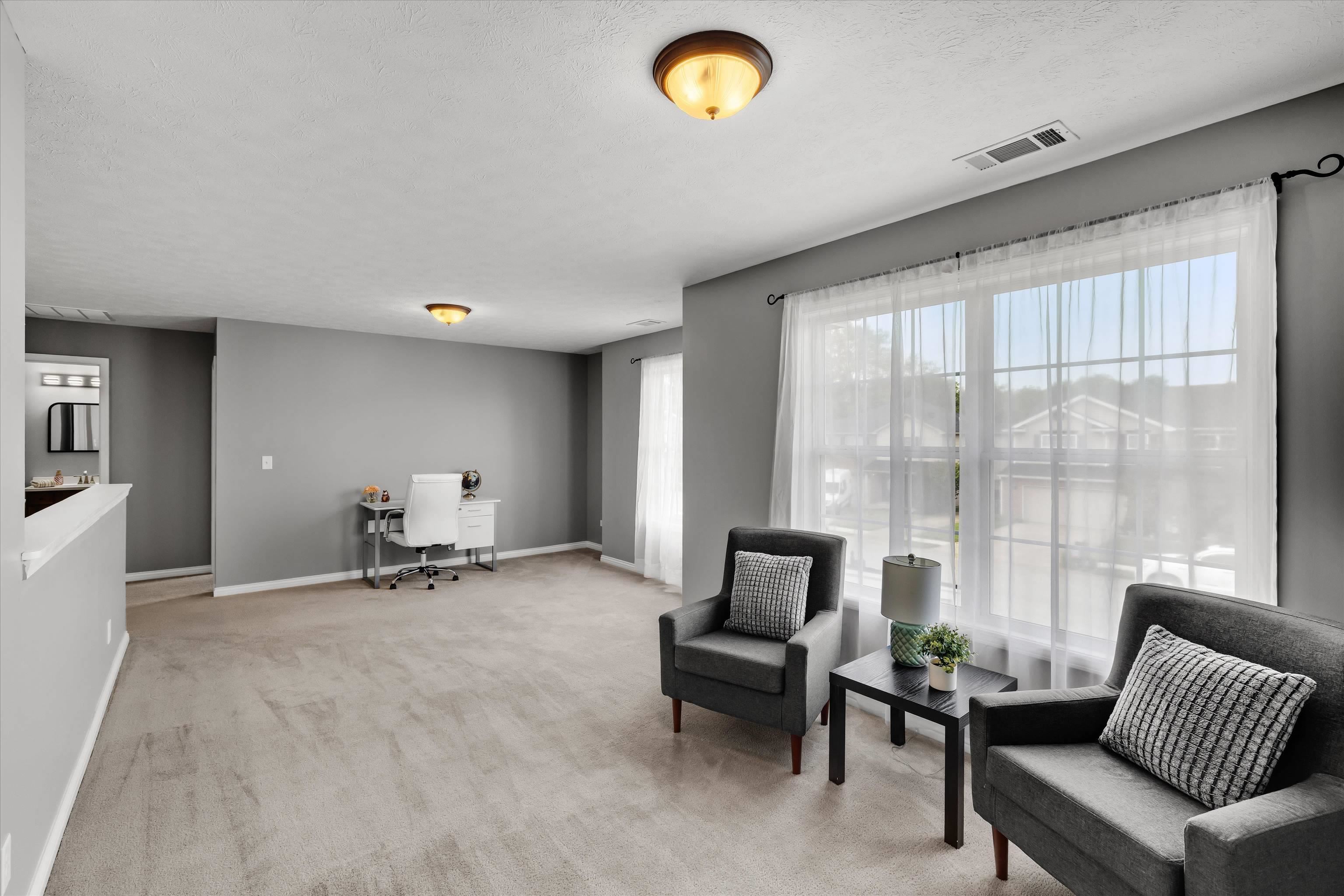 Staged living area with gray armchairs, a desk, and large curtained windows.