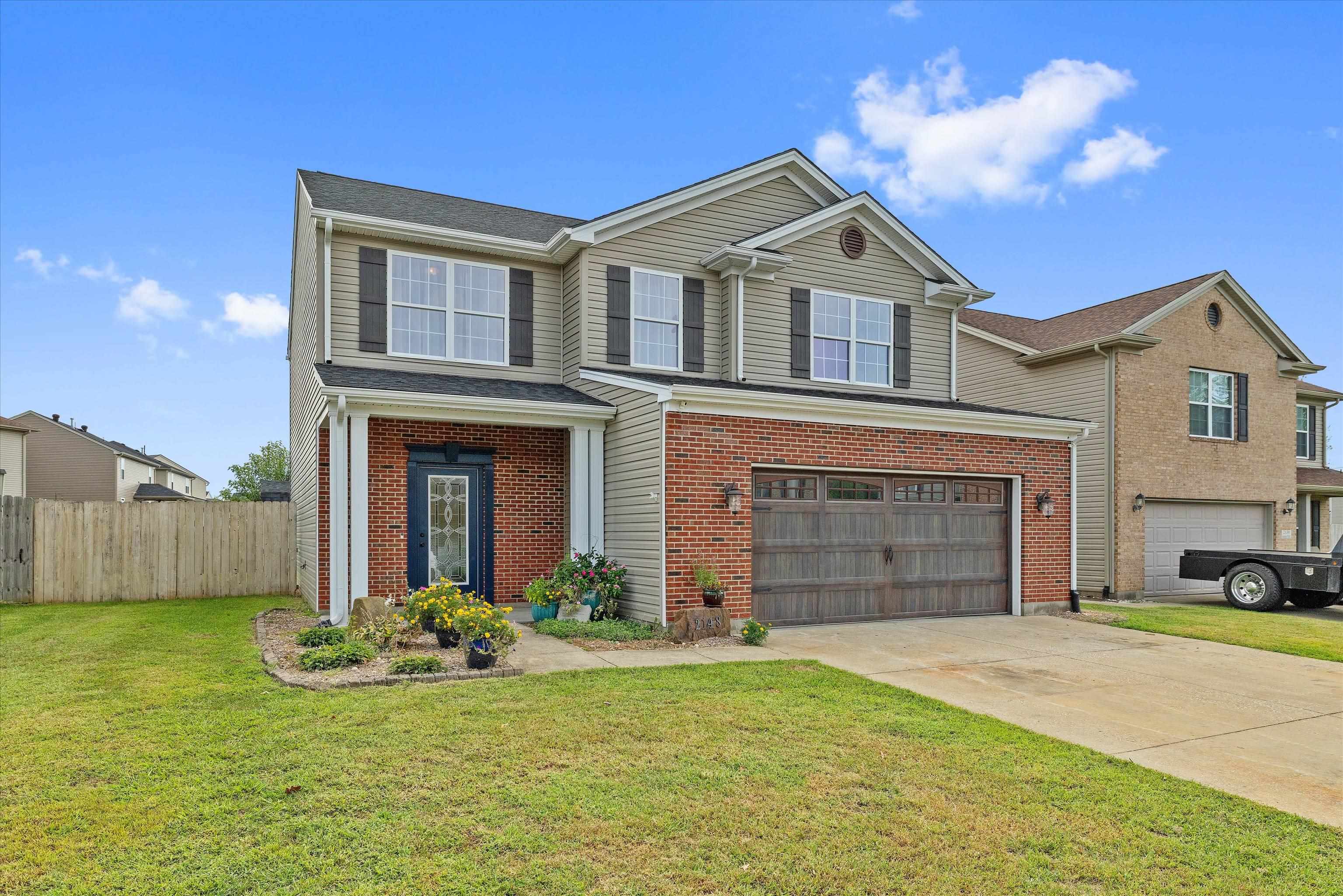 Two-story beige-sided and red-brick home with a blue front door, flower garden, and two-car garage.