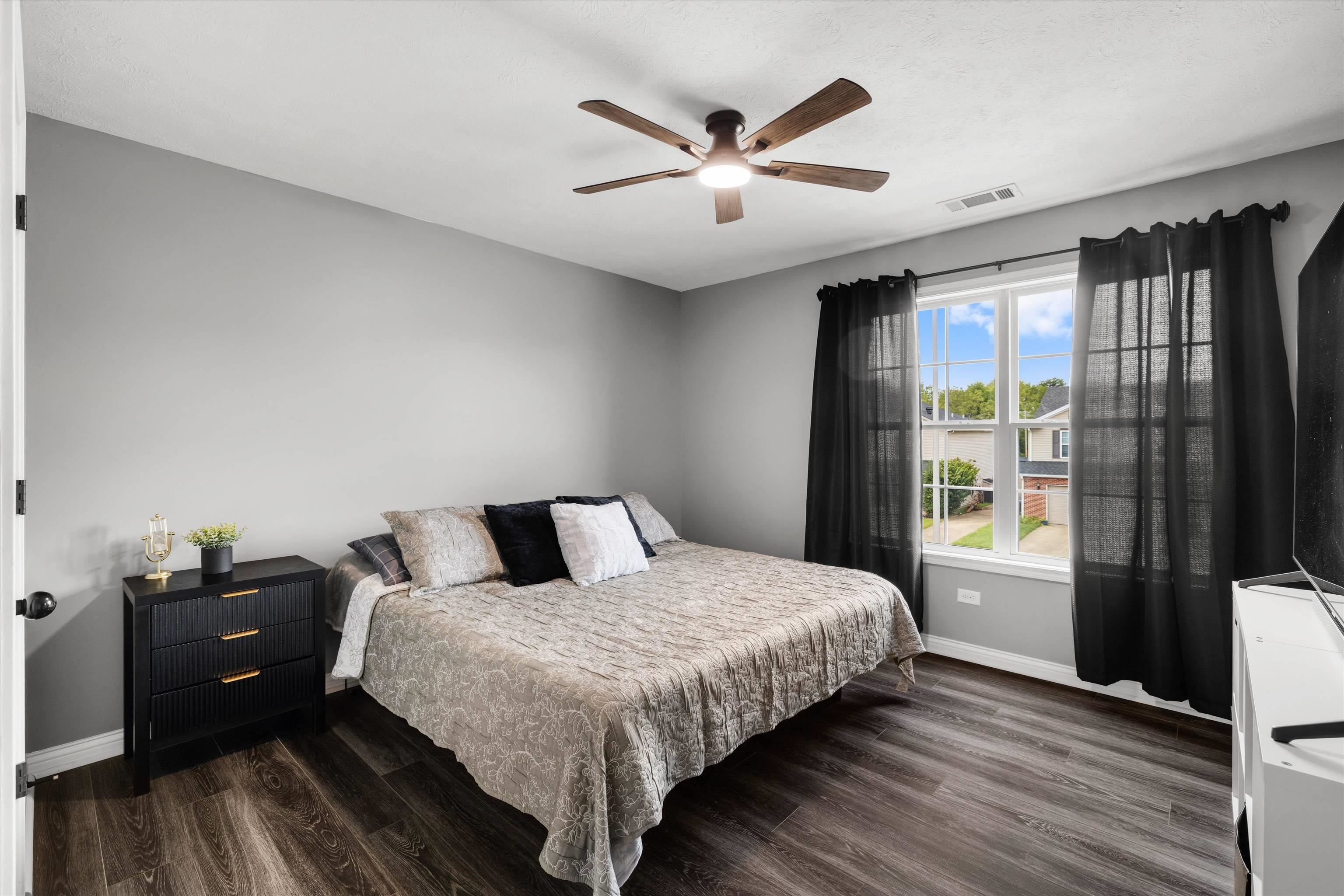 Furnished bedroom with a patterned bed, dark flooring, and black curtains framing a bright window.