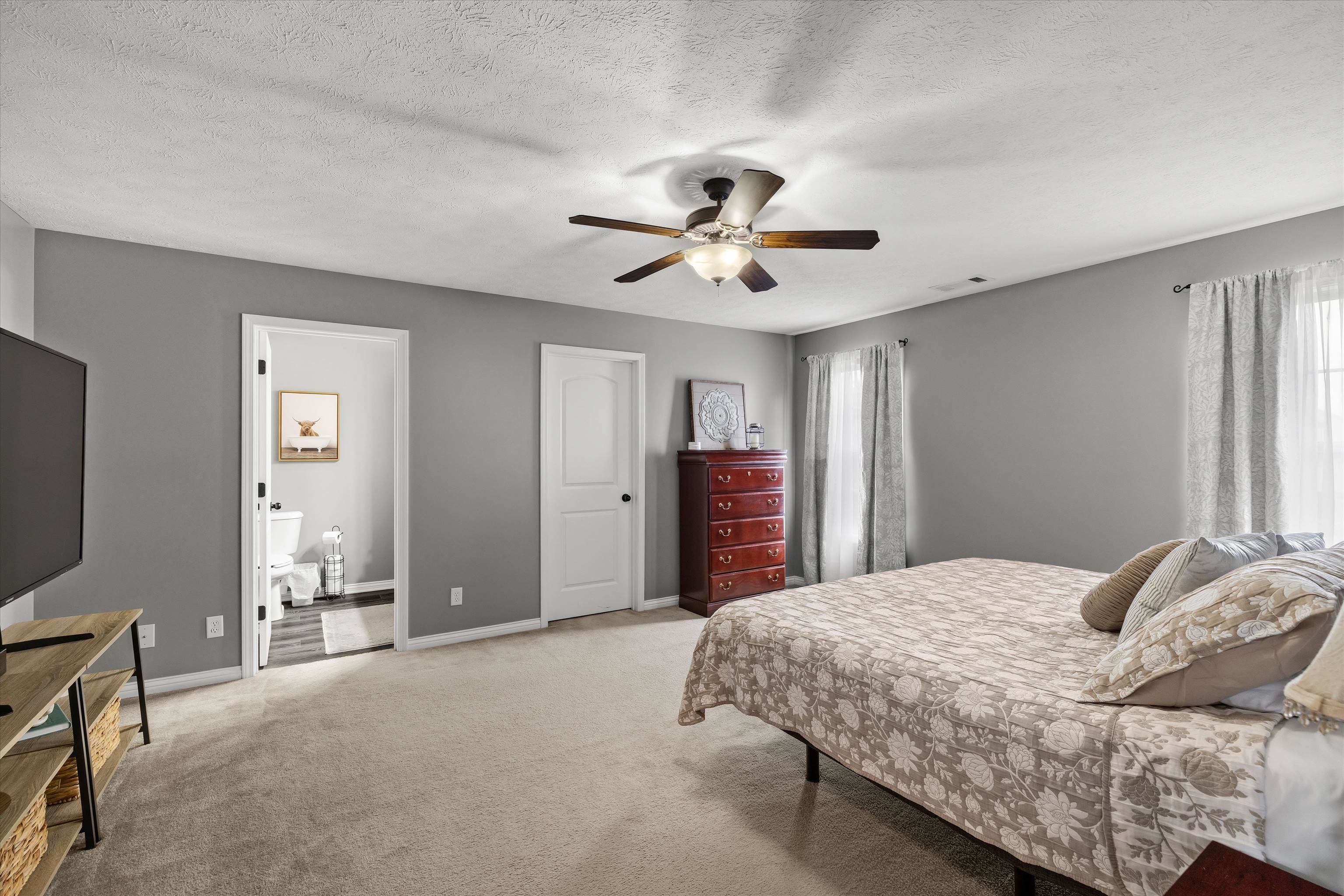 Furnished bedroom with a patterned bedspread, ceiling fan, and open doorway to the bathroom.