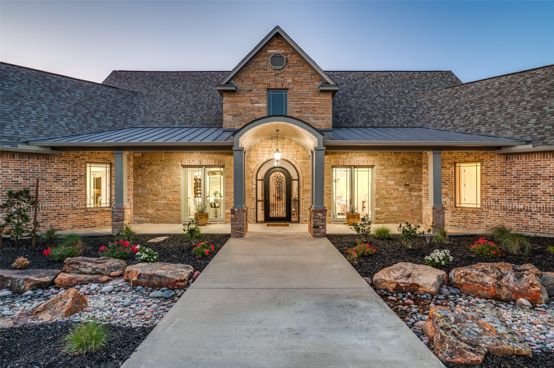 Brick and stone home with an arched entrance, lit windows, and landscaped walkway at dusk.