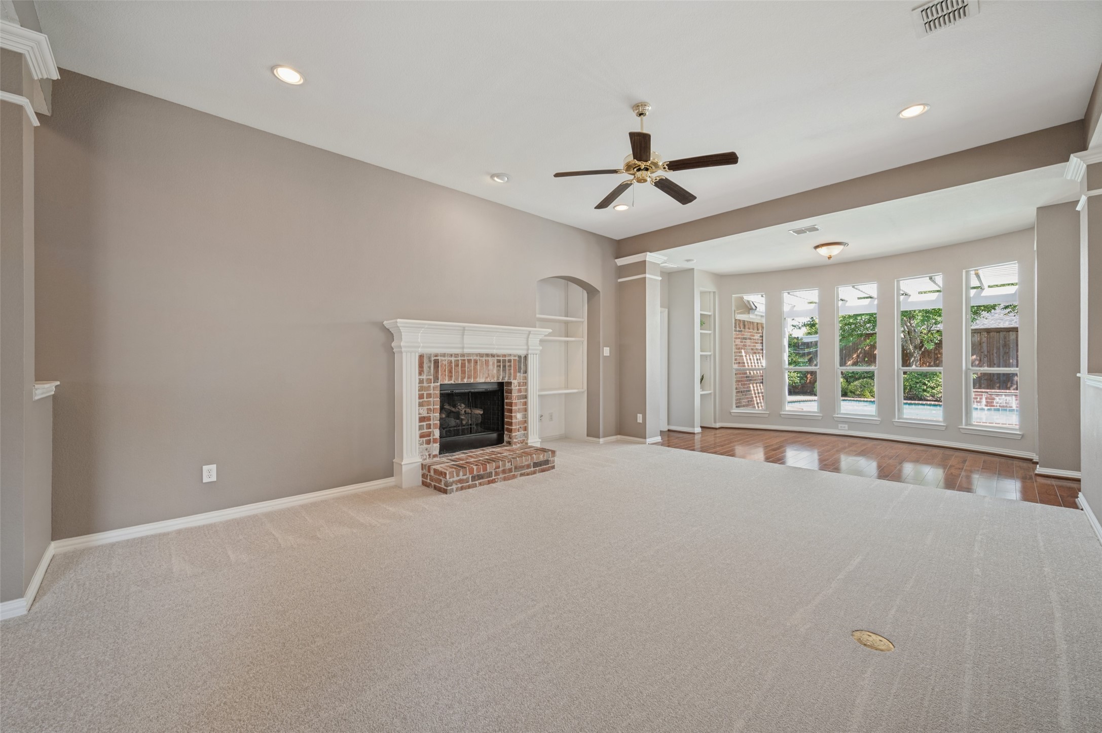 Empty living room with a brick fireplace, beige carpet, and tall windows overlooking a backyard pool.