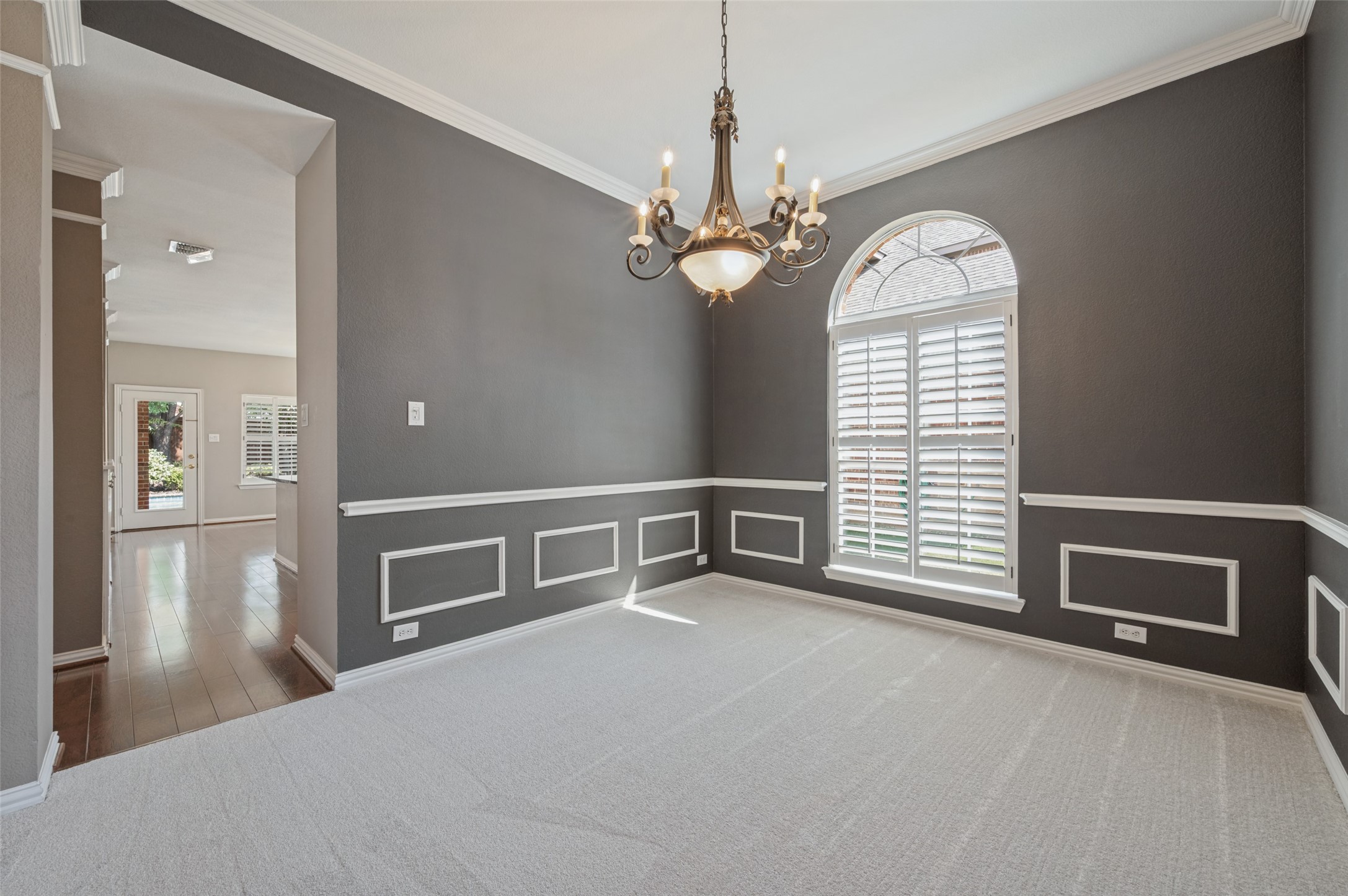 Empty dining room with an ornate chandelier, gray paneled walls, and an arched shuttered window.