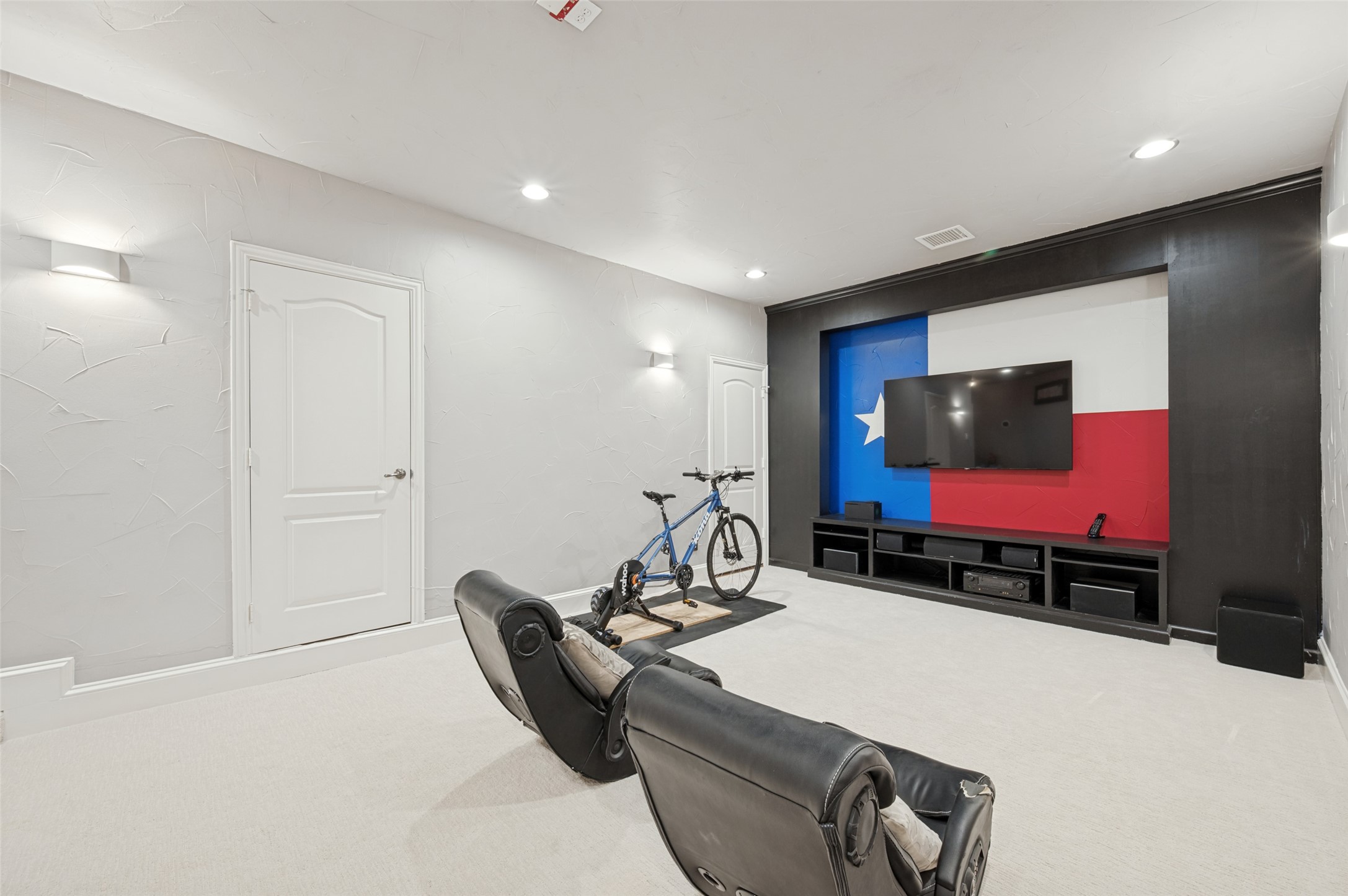 Media room with a Texas flag accent wall, wall-mounted TV, leather recliners, and an indoor bicycle trainer.