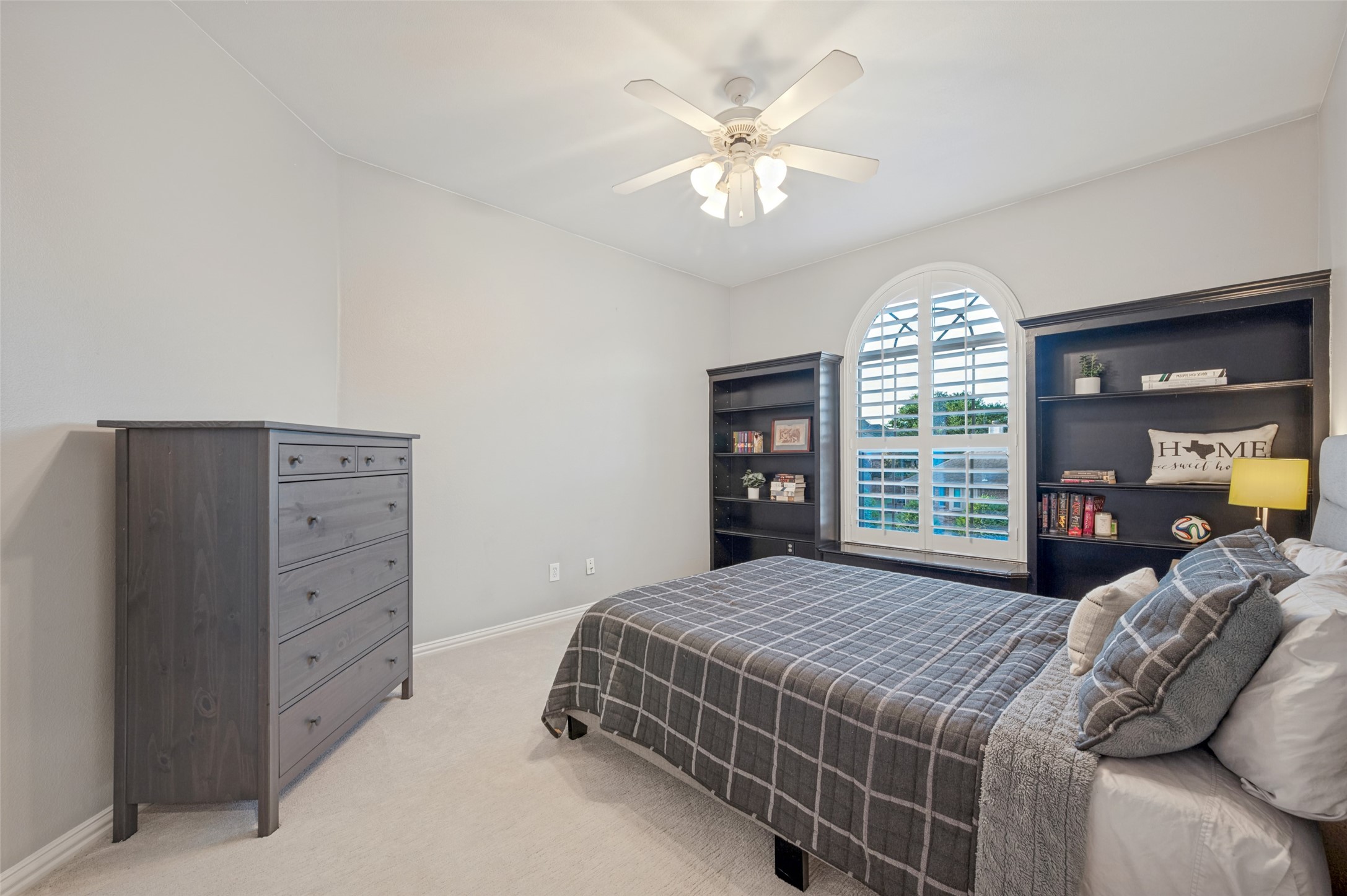 Bedroom with gray plaid bed, dark built-in bookcases, and an arched shuttered window.