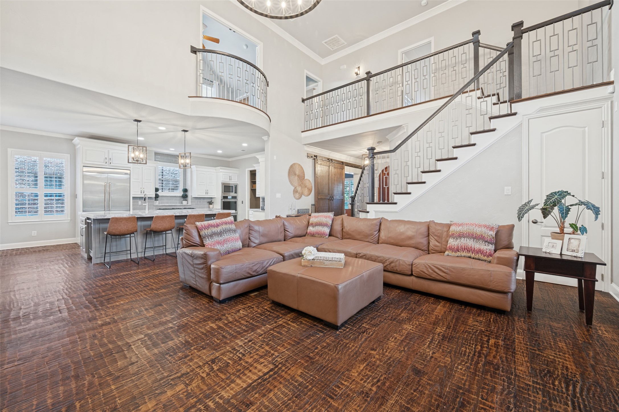 Open-plan living room with leather sectional, dark wood floors, kitchen island, and sweeping staircase.
