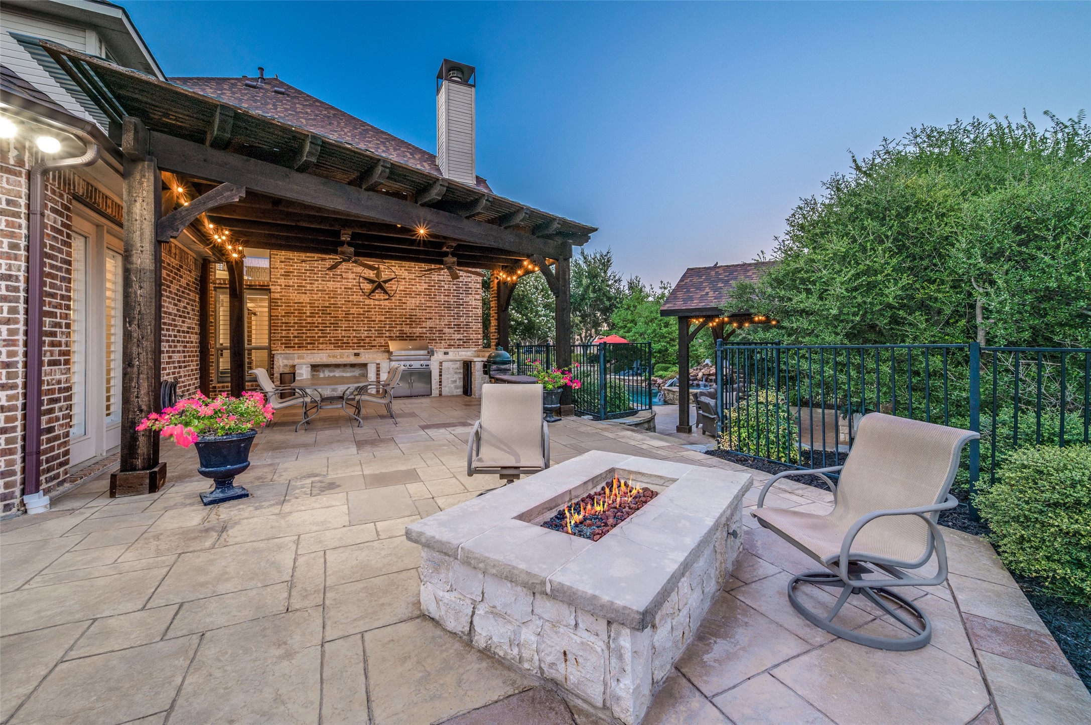 Stone patio with covered outdoor kitchen, seating, a lit fire pit, and pool glimpsed beyond the railing.