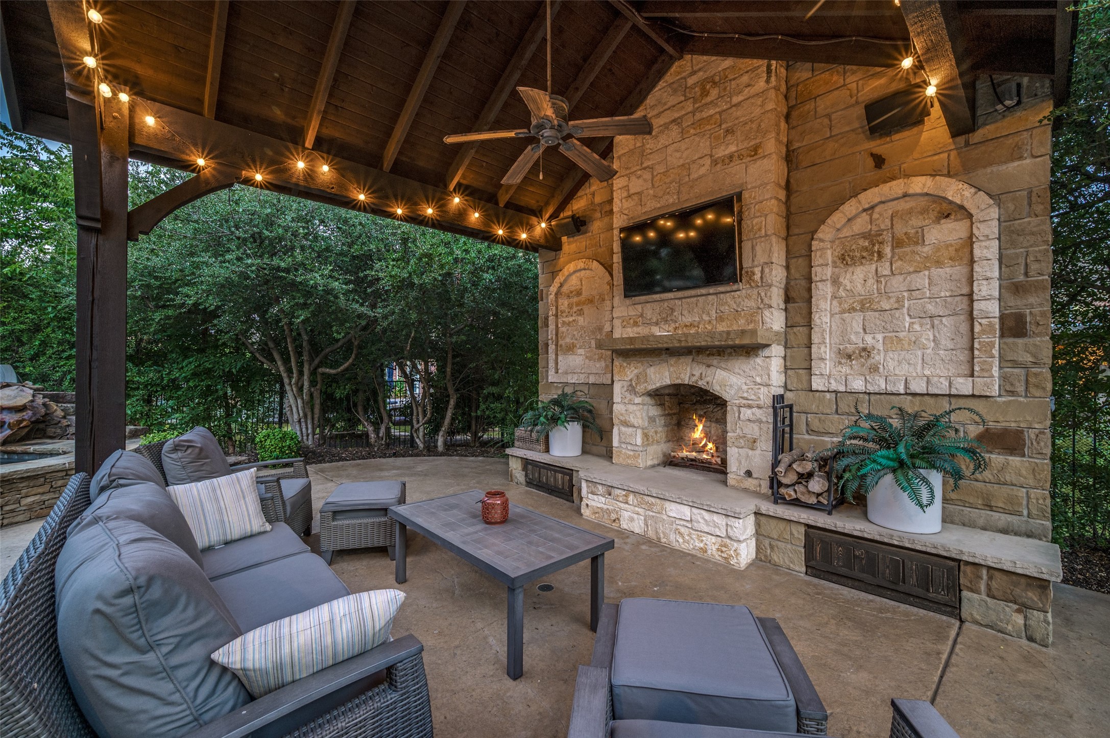 Covered stone patio with fireplace, cushioned seating, and string lights beneath a timber roof.