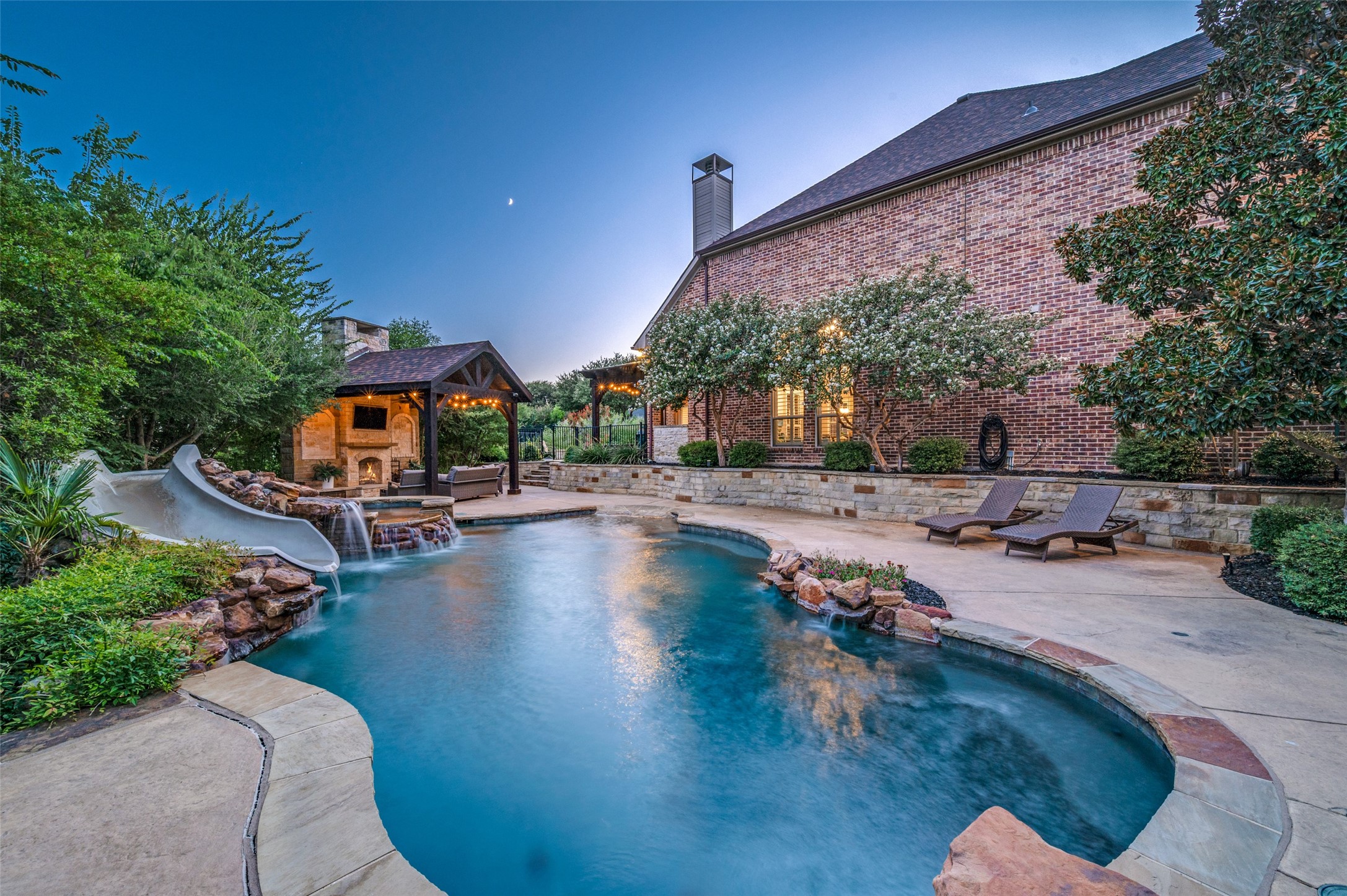 Backyard pool beside a brick home, with a waterslide, rock waterfall, and illuminated fireplace pavilion.