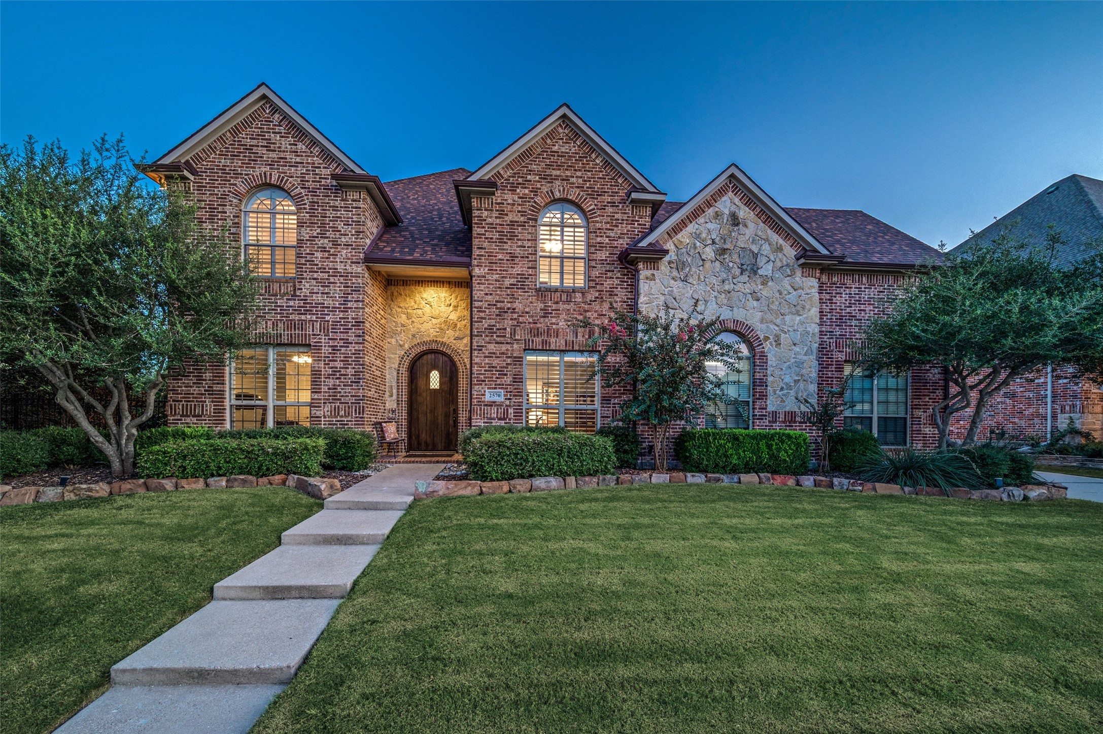 Two-story brick and stone home with illuminated windows and a walkway to the arched entry.