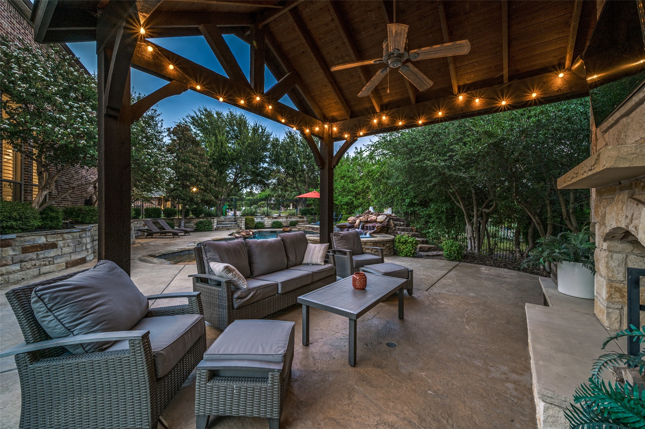Covered patio with wicker seating and string lights overlooking a landscaped pool and rock waterfall.