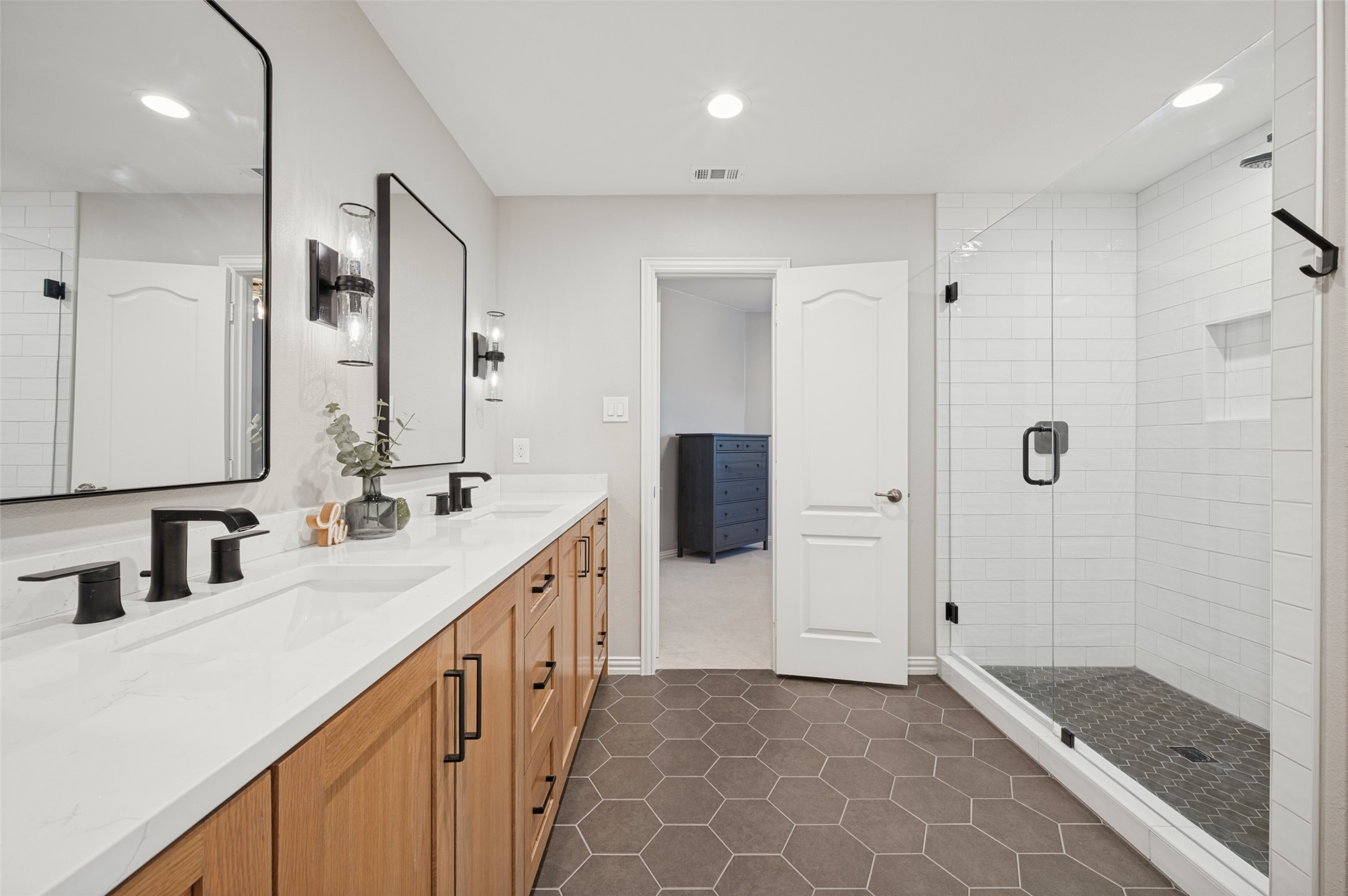 Bathroom with double wood vanity and glass walk-in shower tiled in white and dark hexagons.