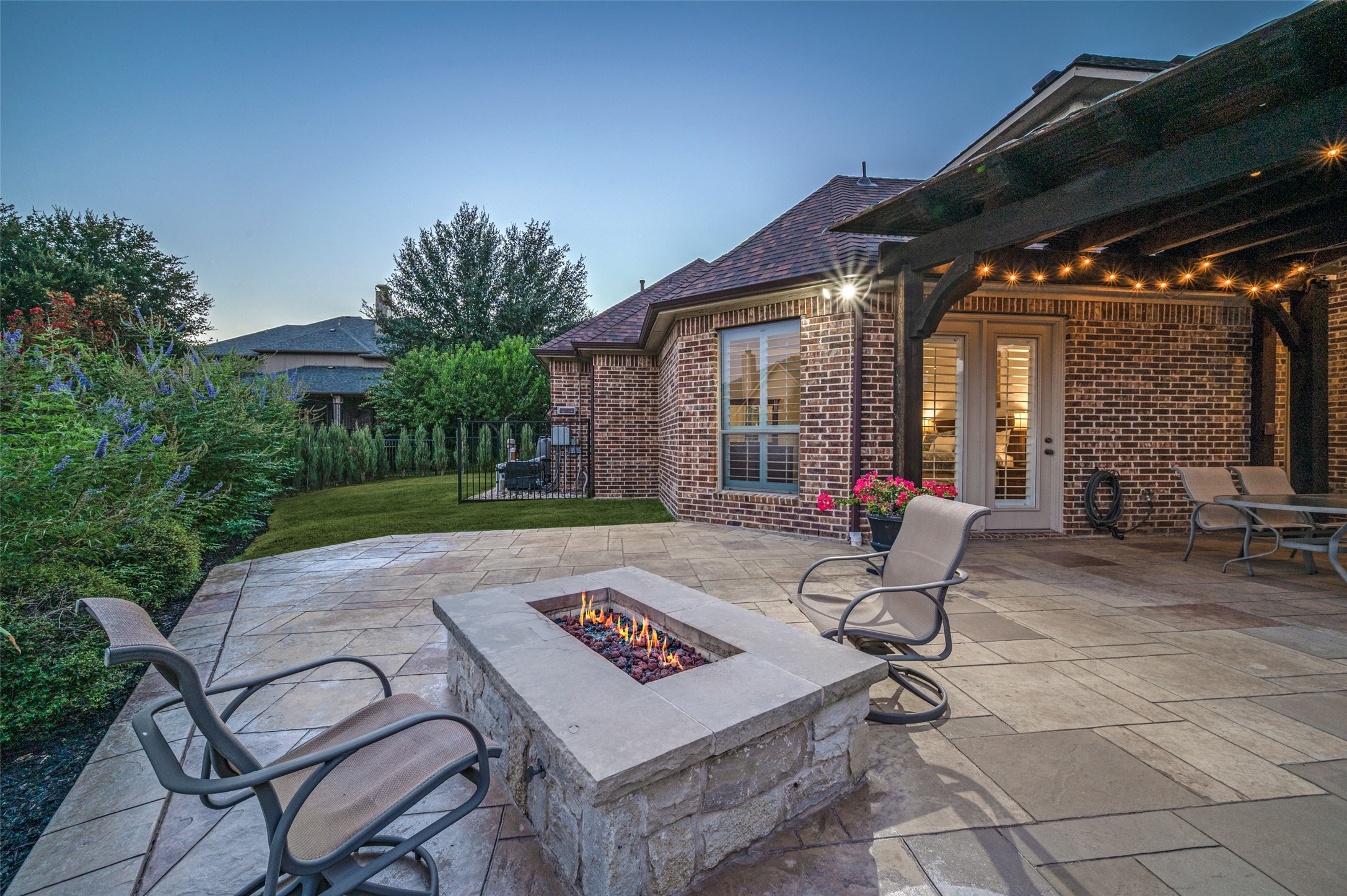 Backyard patio with a stone fire pit, covered pergola, and glowing string lights at dusk.