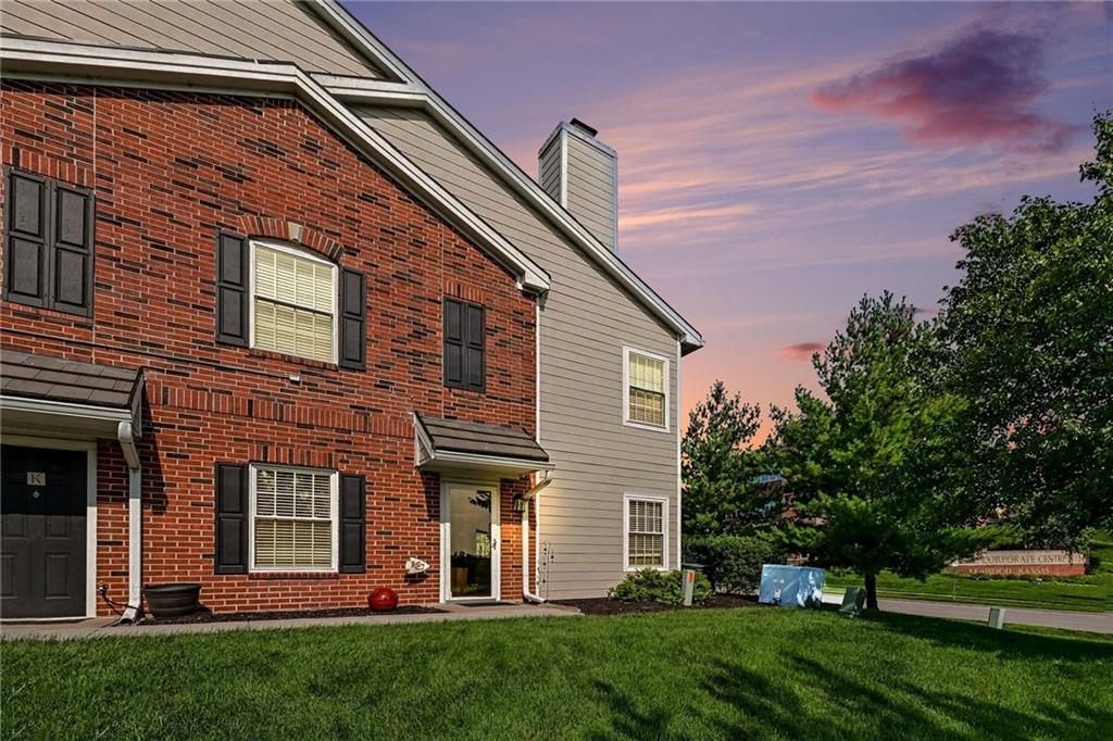 Two-story brick-and-siding townhouse with black shutters, a green lawn, and a pink dusk sky.