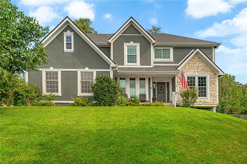 Gray two-story house with stone facade, covered porch, American flag, and broad green lawn.