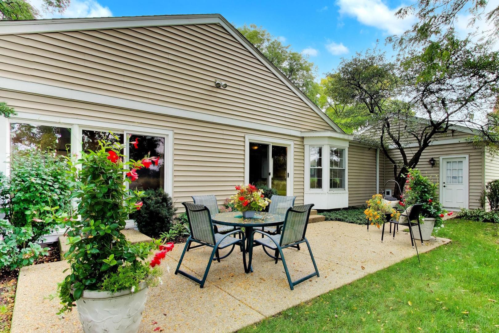 Home’s rear patio with outdoor dining furniture and flowering planters beside beige siding.