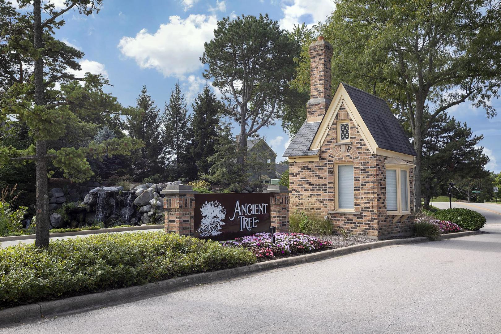 Ancient Tree community entrance with a brick sign, gabled guardhouse, stone waterfall, and flower beds.