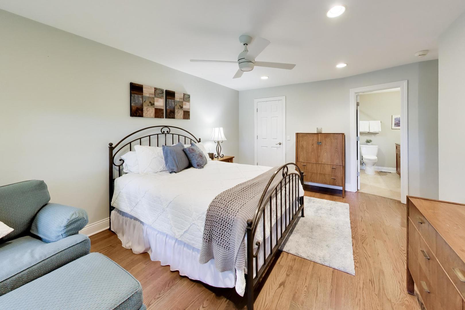 Staged bedroom with a white bed, dark metal frame, blue chair, and open bathroom doorway.