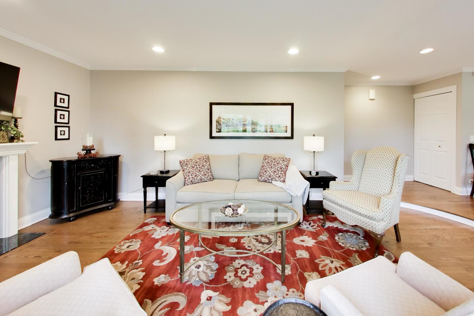 Living room with a light sofa, patterned armchair, and red floral rug on hardwood floors.