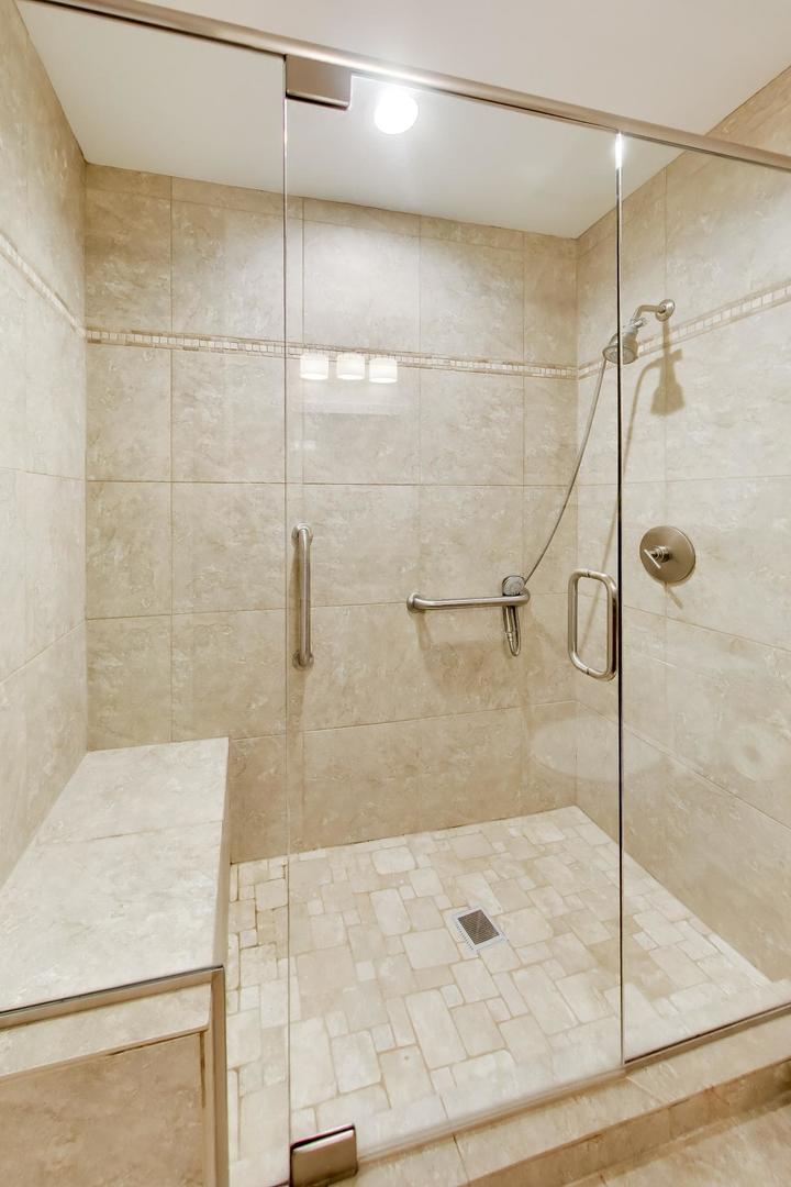 Walk-in shower with beige stone tiles, clear glass enclosure, grab bars, and a built-in bench.