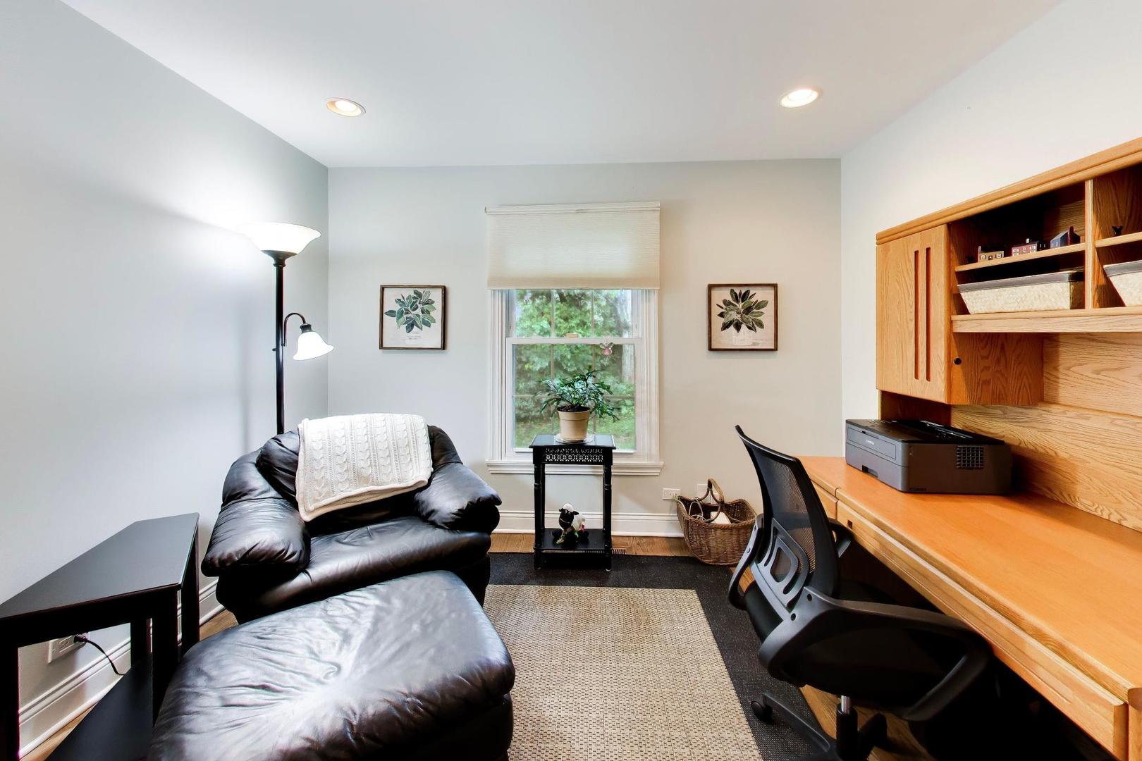 Home office with oak built-in desk, black chair, and leather recliner beside a window.