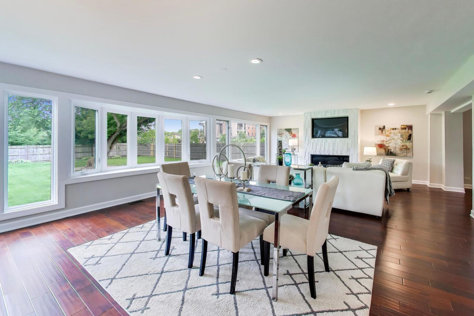 Open-plan dining and living room with a fireplace, mounted television, and windows overlooking a fenced yard.