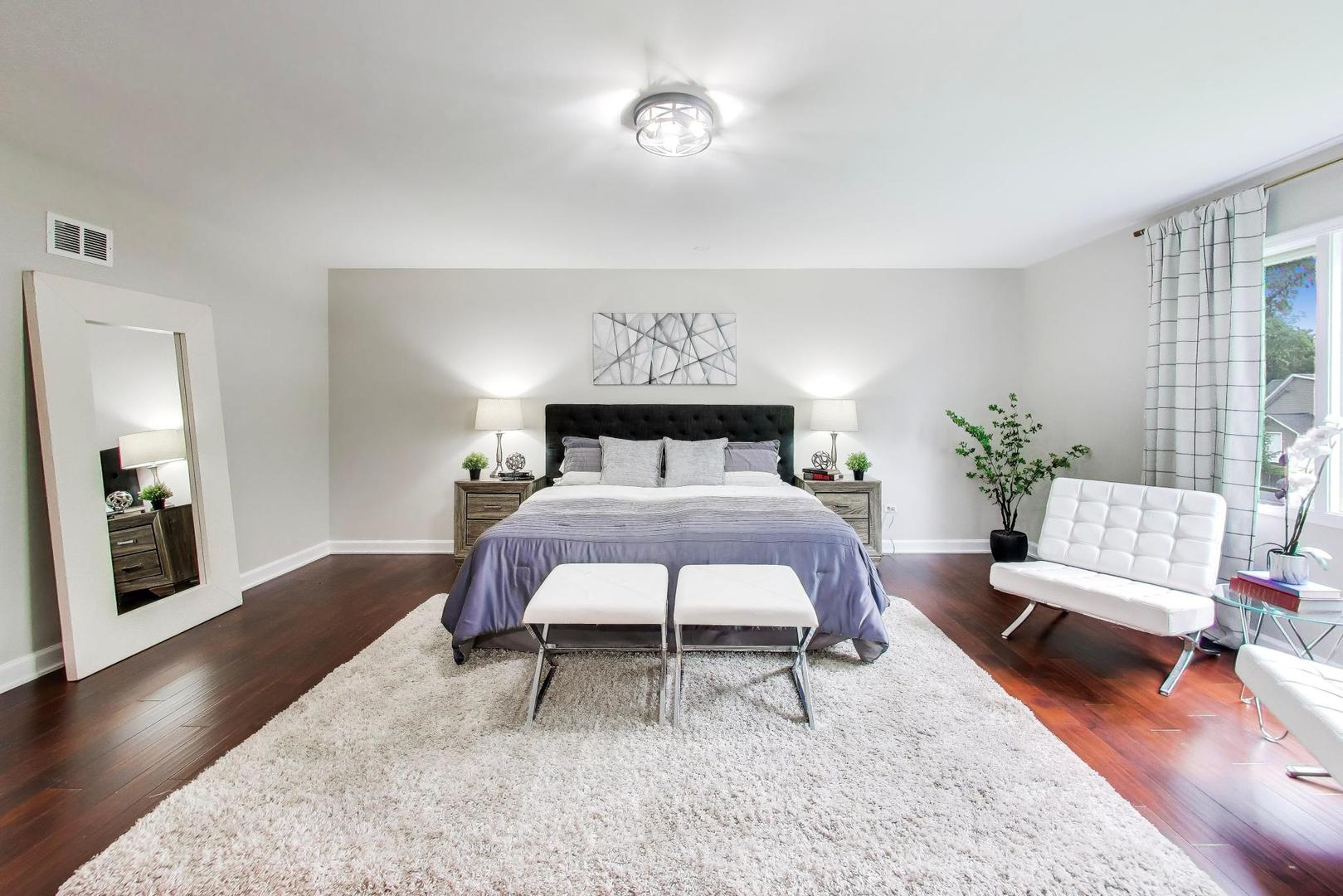Staged bedroom with a tufted dark headboard, white rug, and tall window.