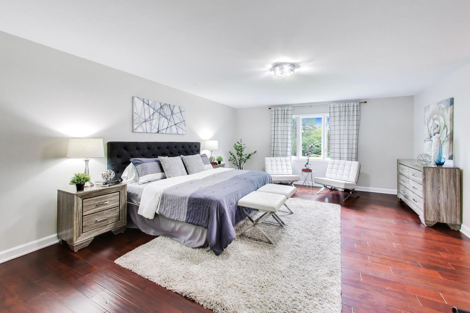 Staged bedroom with a gray bed, white window-side chairs, dark hardwood floors, and a cream rug.