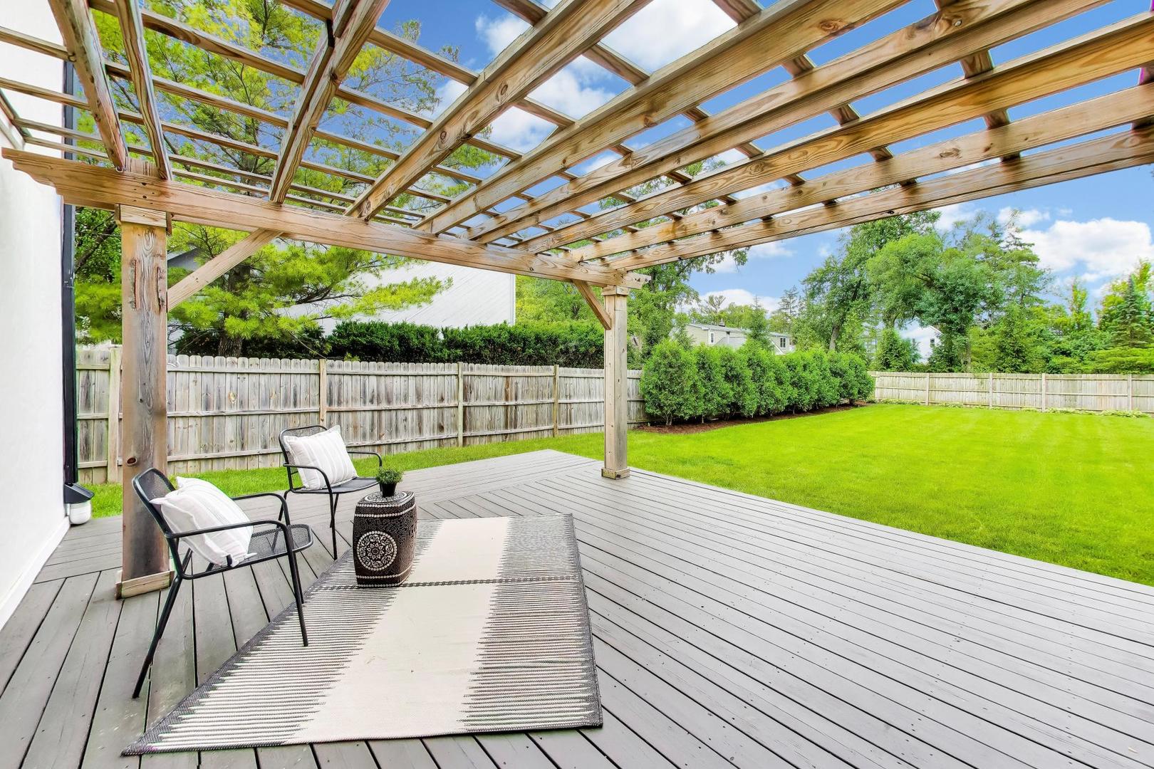 Furnished deck beneath a wooden pergola, overlooking a fenced lawn.