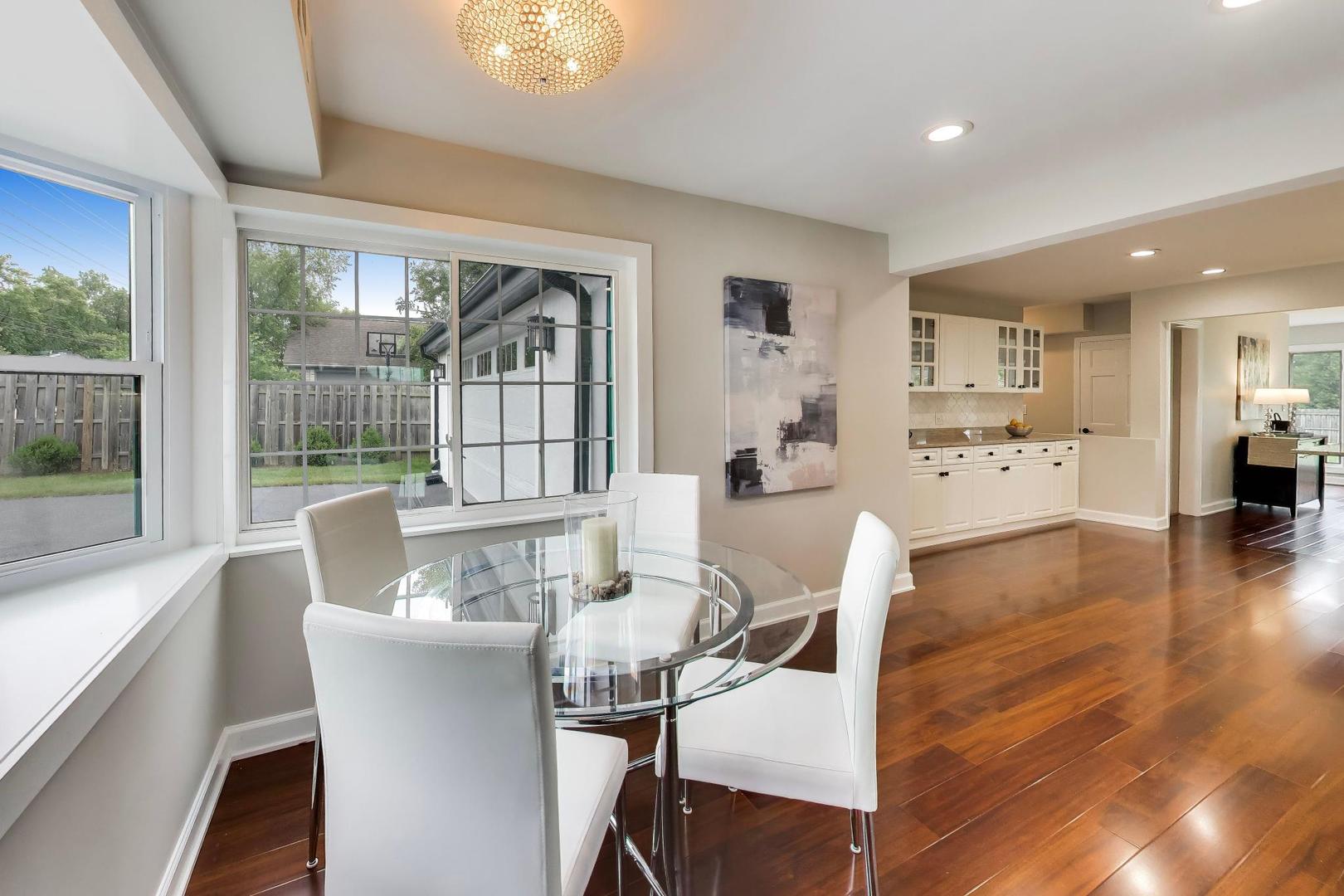 Dining area with a round glass table beside large windows and an open white-cabinet kitchen.