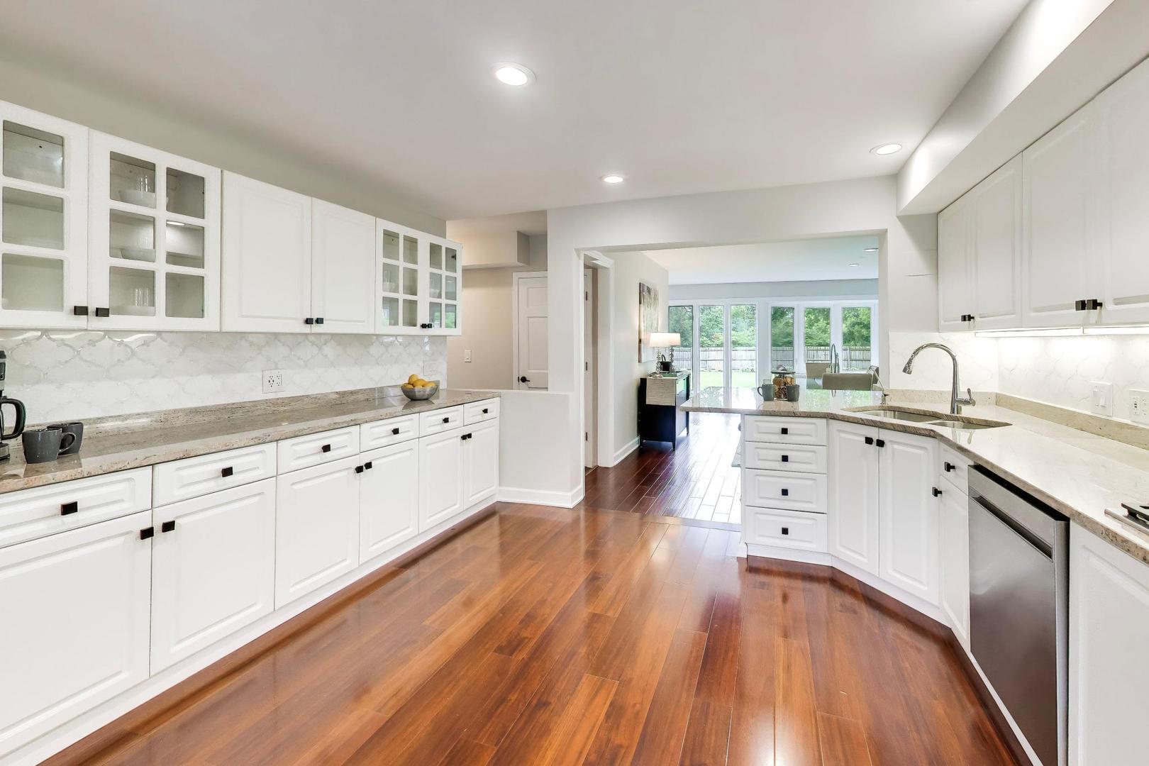 White kitchen with glass-front cabinets, stone counters, and wood floors opening to a bright dining area.
