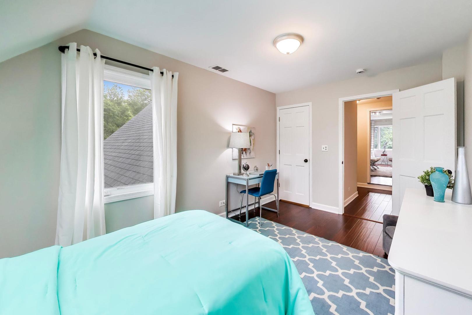 Furnished bedroom with a turquoise bedspread, curtained window, and blue desk chair.