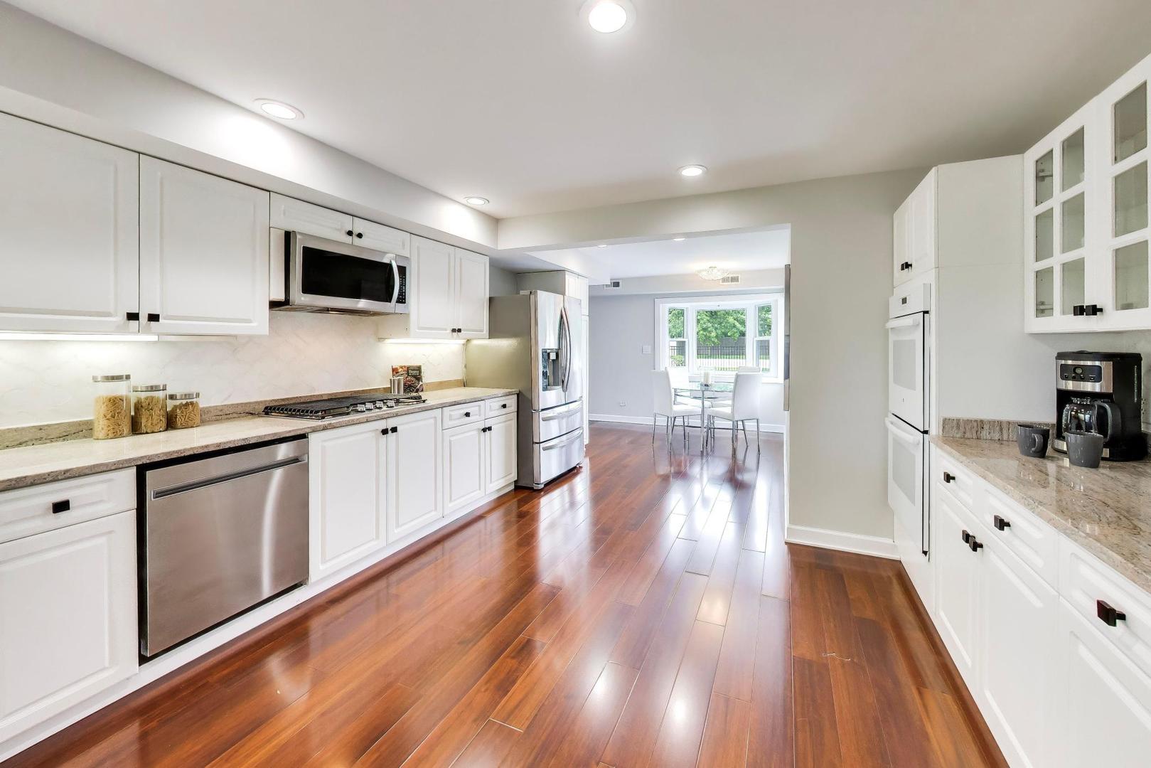 Bright white kitchen with stainless steel appliances, stone counters, and hardwood floors opening to a dining area.