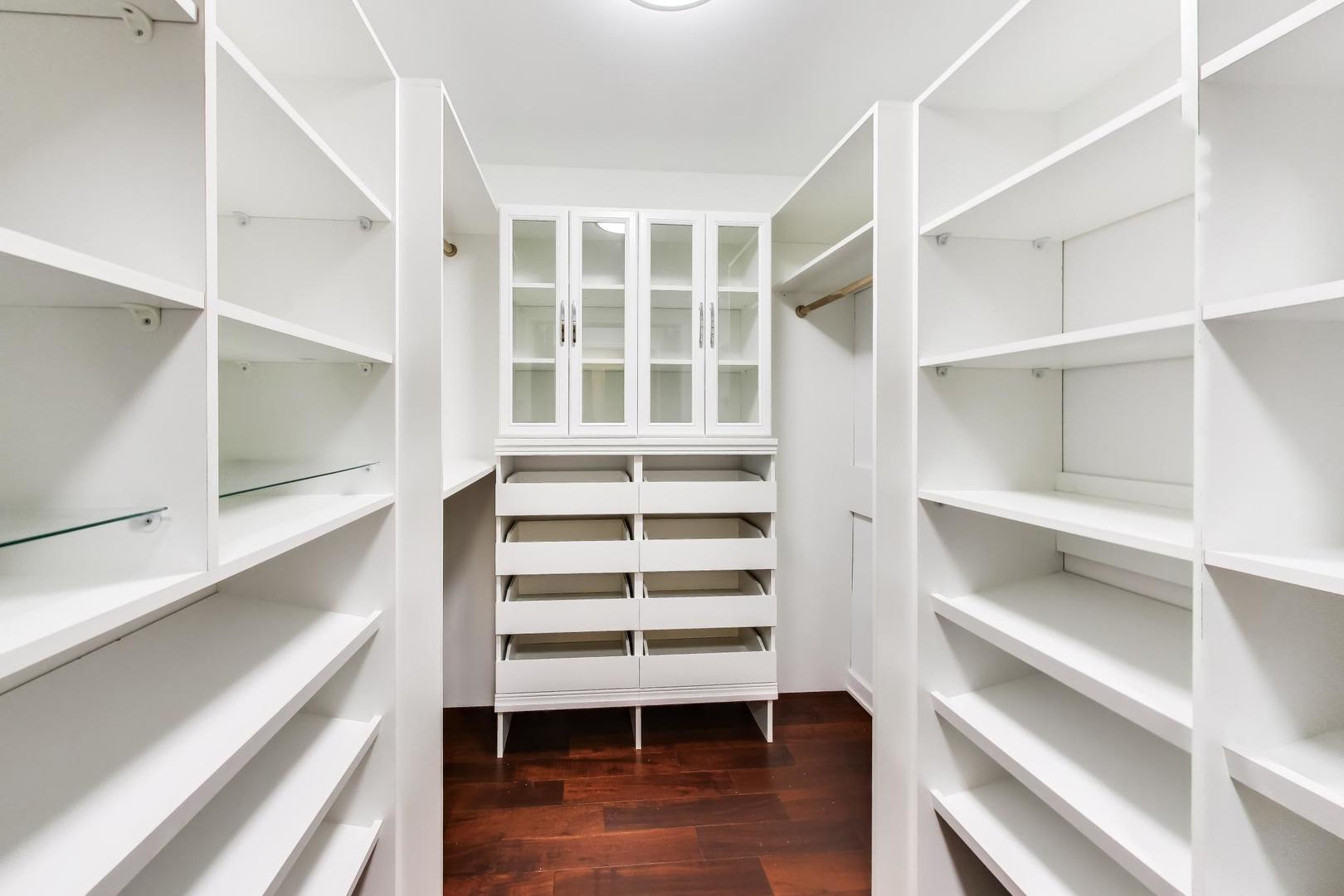 Walk-in closet with white built-in shelves, central drawers, glass-front cabinets, and dark hardwood floors.