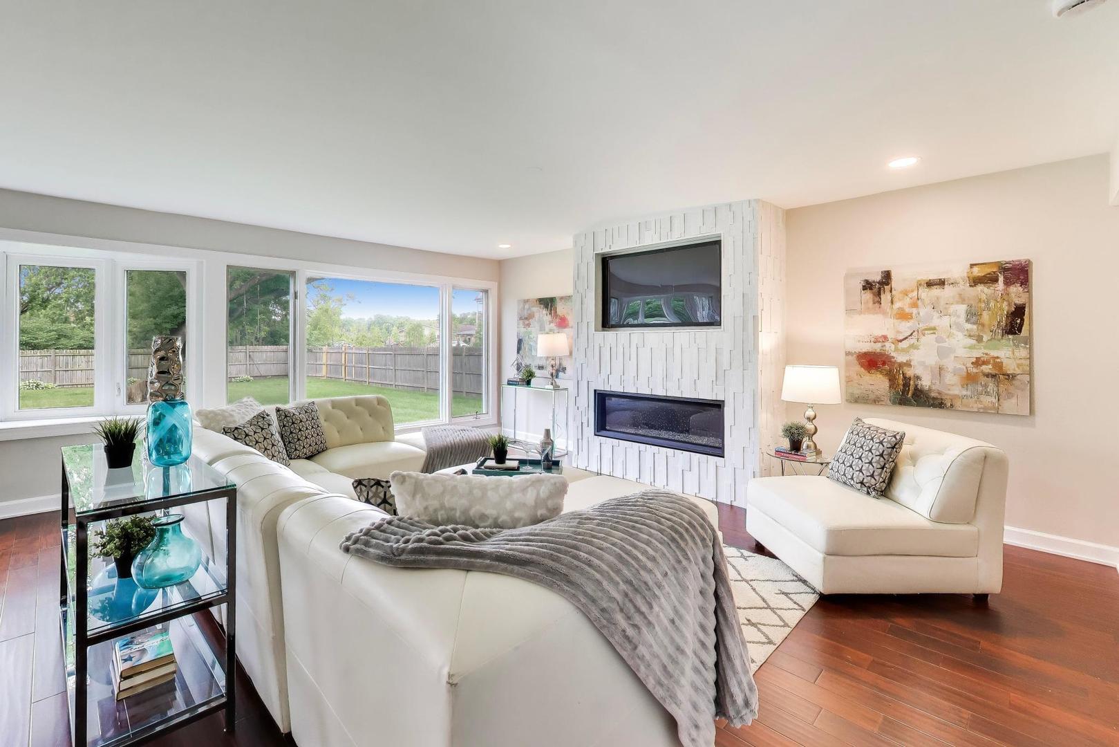 Living room with white sofas, a modern fireplace, and large windows overlooking a fenced backyard.