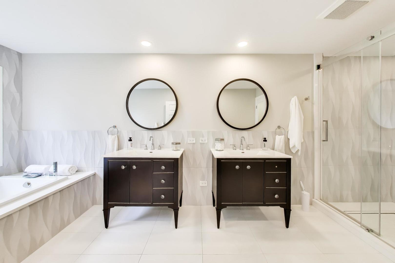 Bathroom with double dark wood vanities, round mirrors, a soaking tub, and glass shower.