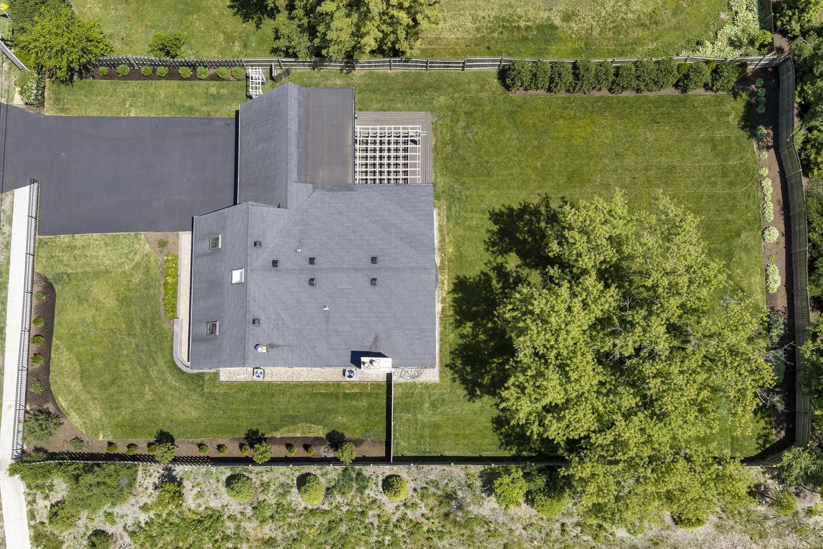 Aerial view of a gray-roofed home, fenced lawn, mature trees, and paved driveway.