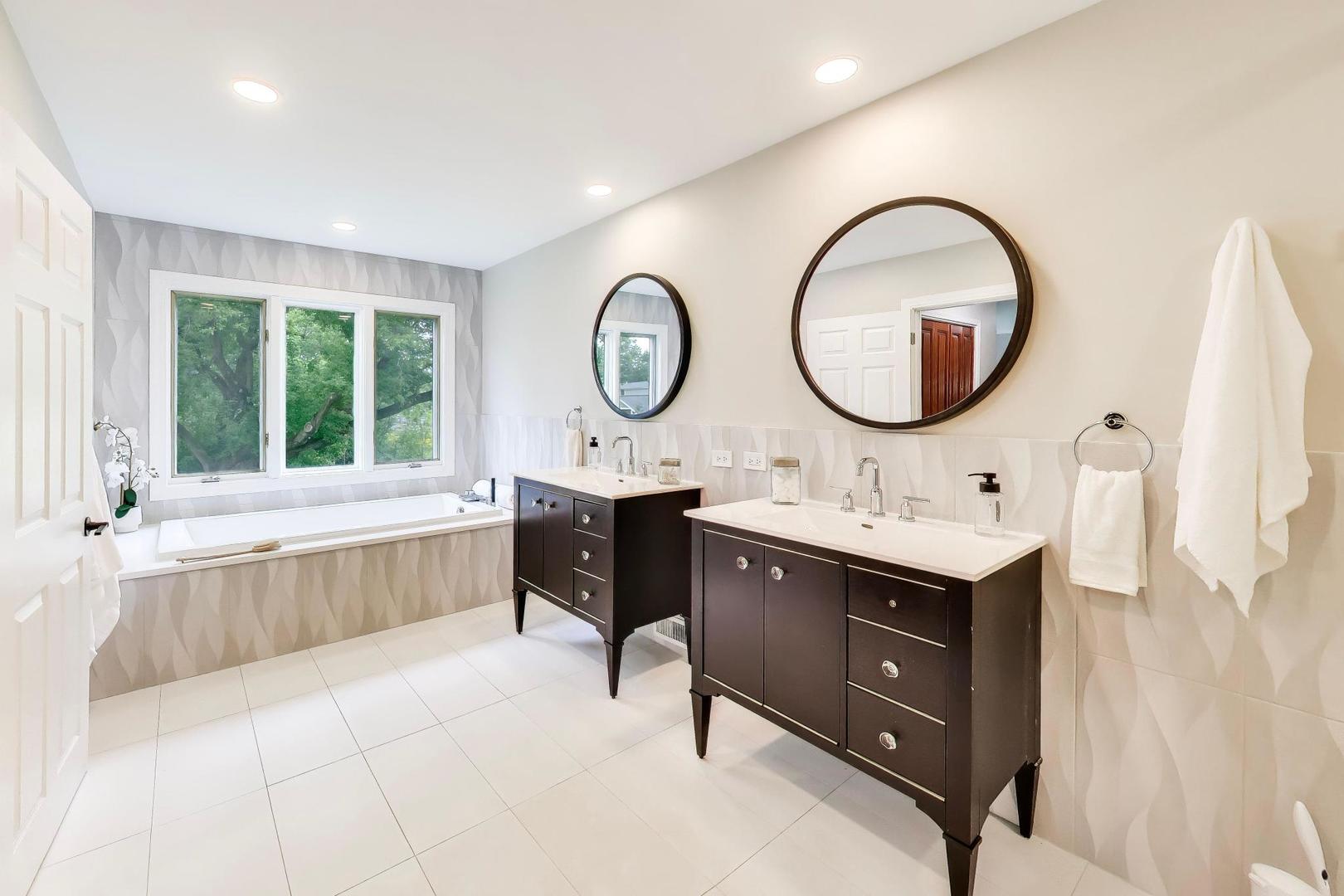 Bathroom with a window-side soaking tub and two dark wood vanities with round mirrors.