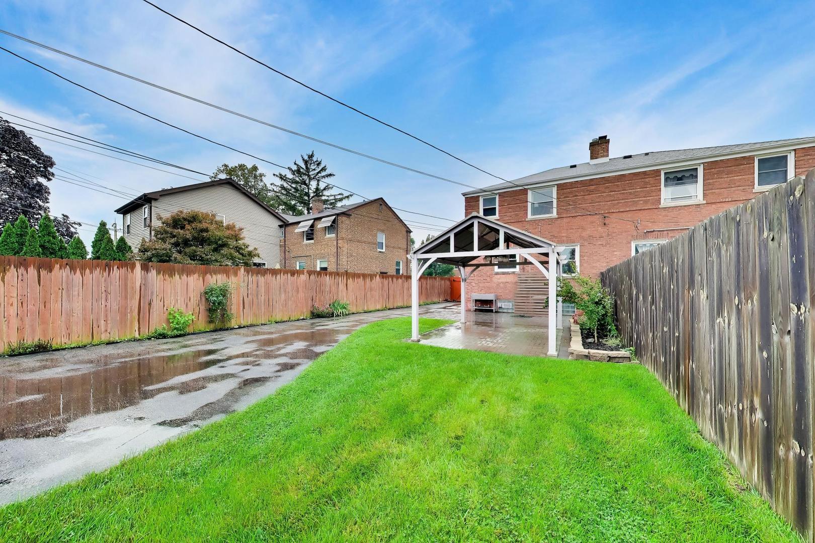 Fenced backyard with a green lawn, covered patio, and wet paved driveway behind a brick house.