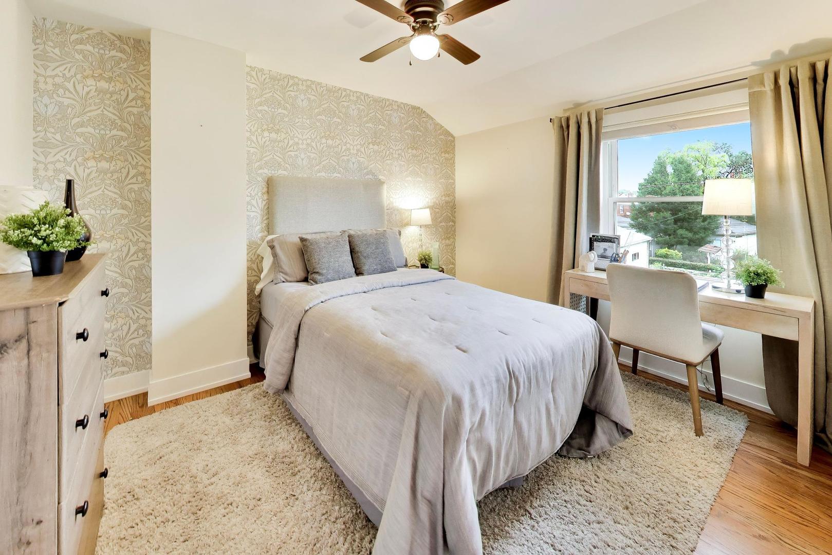 Staged bedroom with a floral accent wall, ceiling fan, and desk beside a large window.