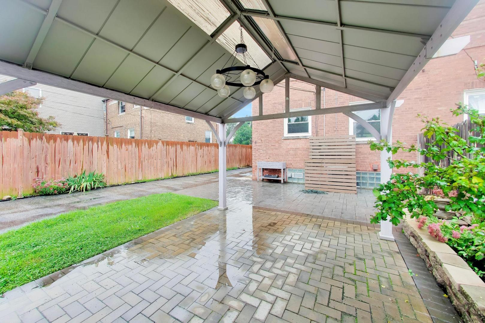 Covered backyard patio with a peaked roof, chandelier, brick pavers, and fenced lawn.