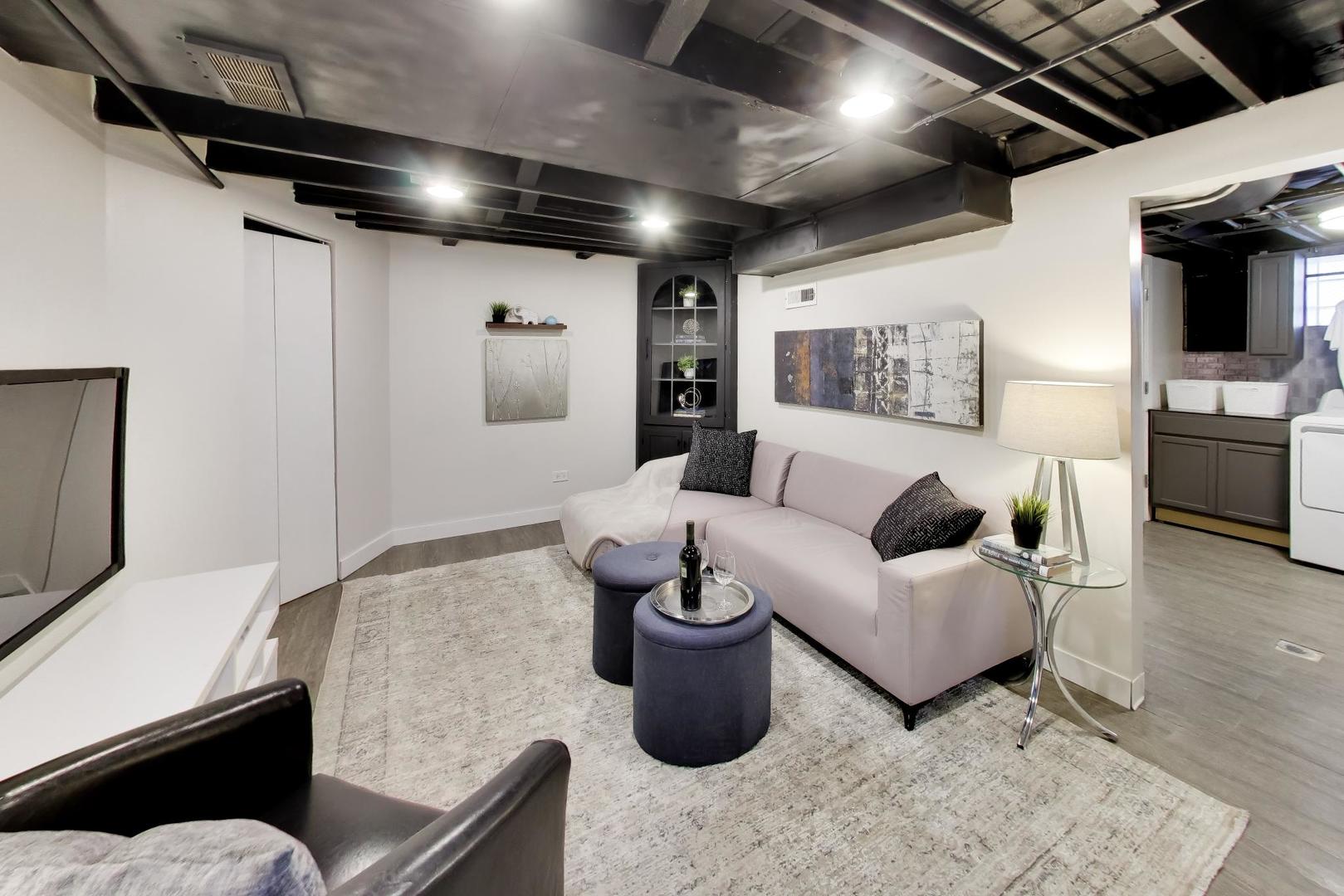 Basement living room with a pale sectional, dark ottomans, exposed ceiling beams, and laundry area doorway.