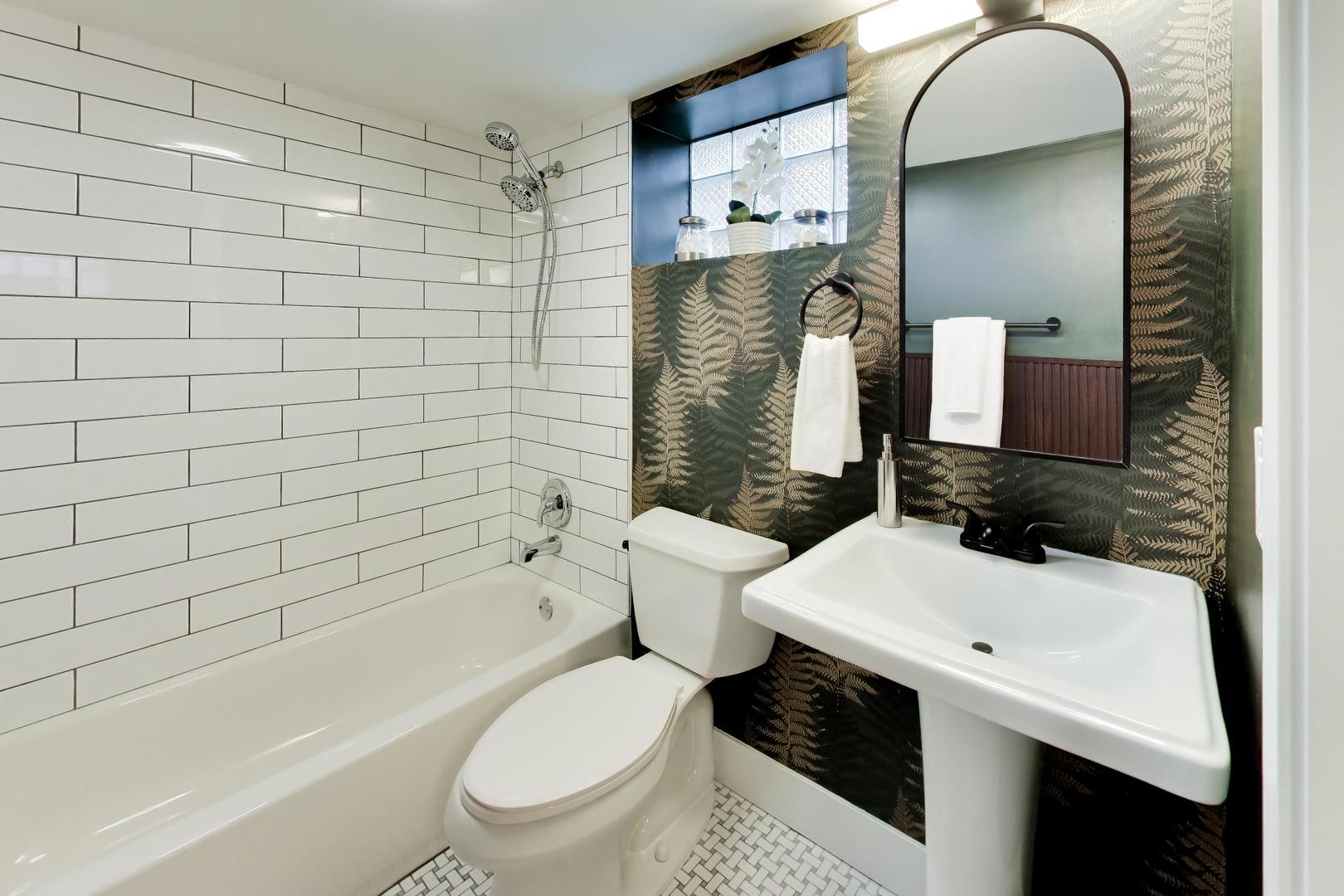 Bathroom with a white subway-tile tub surround, pedestal sink, and dark fern-patterned wallpaper.
