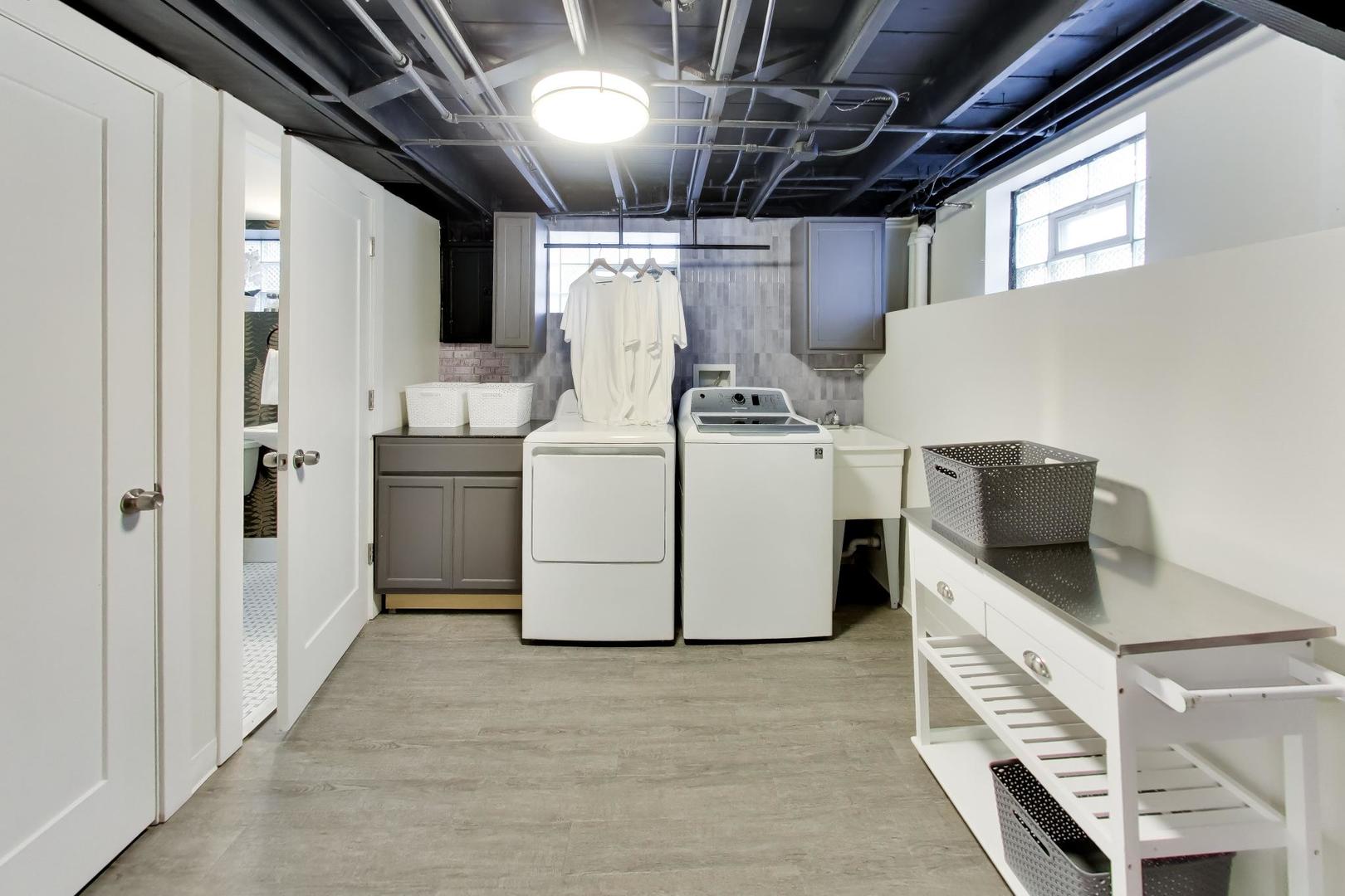 Laundry room with washer and dryer, utility sink, gray cabinets, and a folding table with baskets.