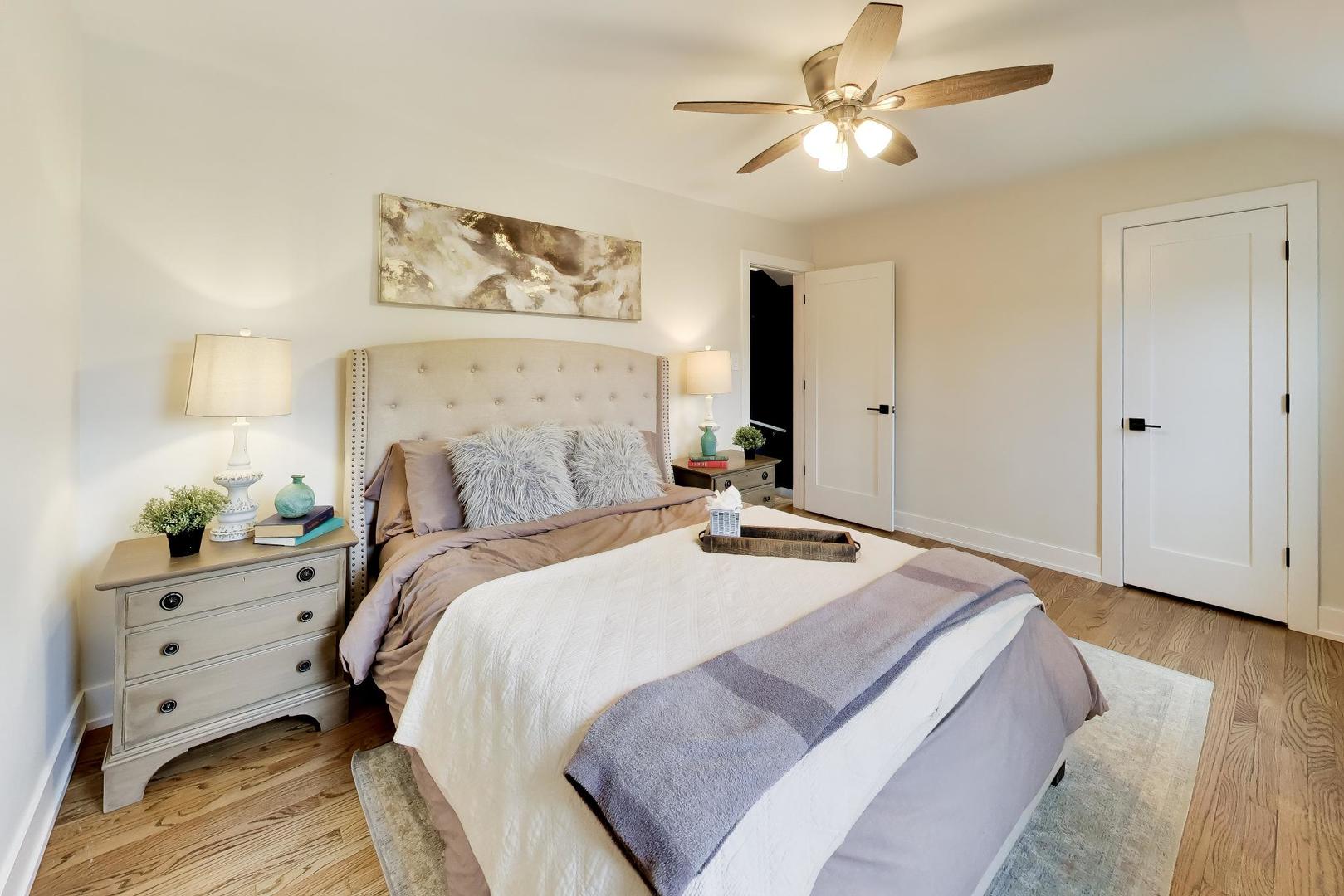 Staged bedroom with a tufted bed, neutral bedding, hardwood floors, and a ceiling fan.