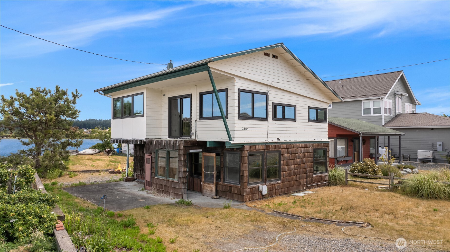 Raised waterfront house with white siding, brown shingle lower level, and water beside the yard.
