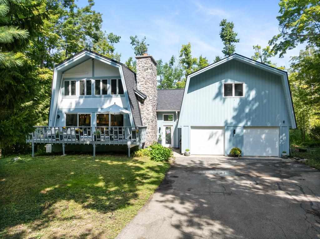 Light blue home with a stone chimney, raised deck, and attached garage among mature trees.
