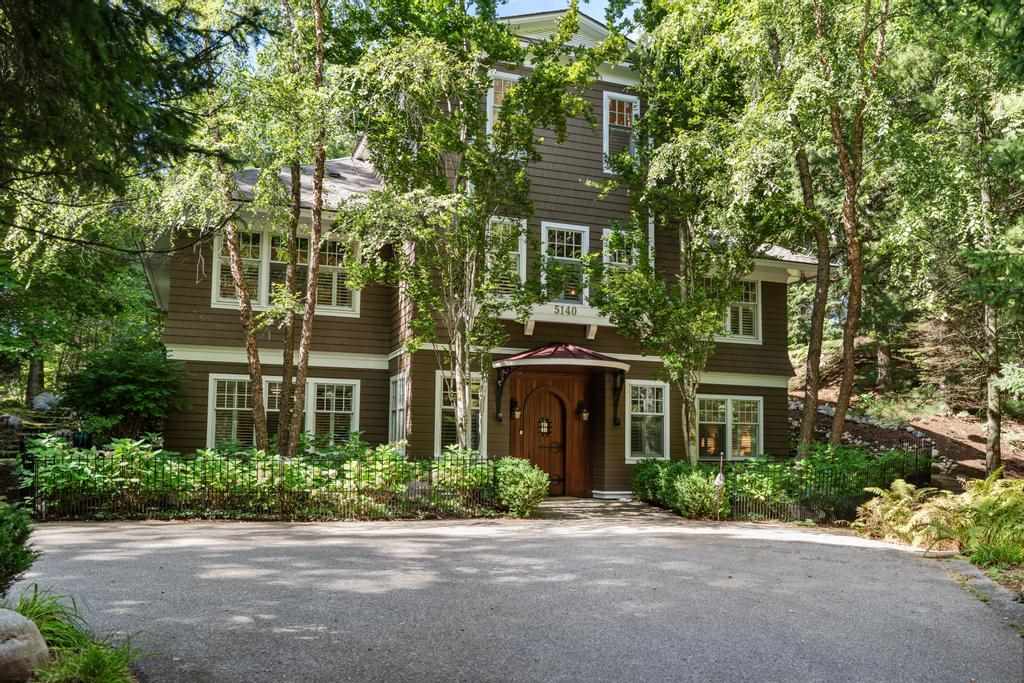 Brown two-story home nestled among mature trees, with a wooden entry door and paved driveway.