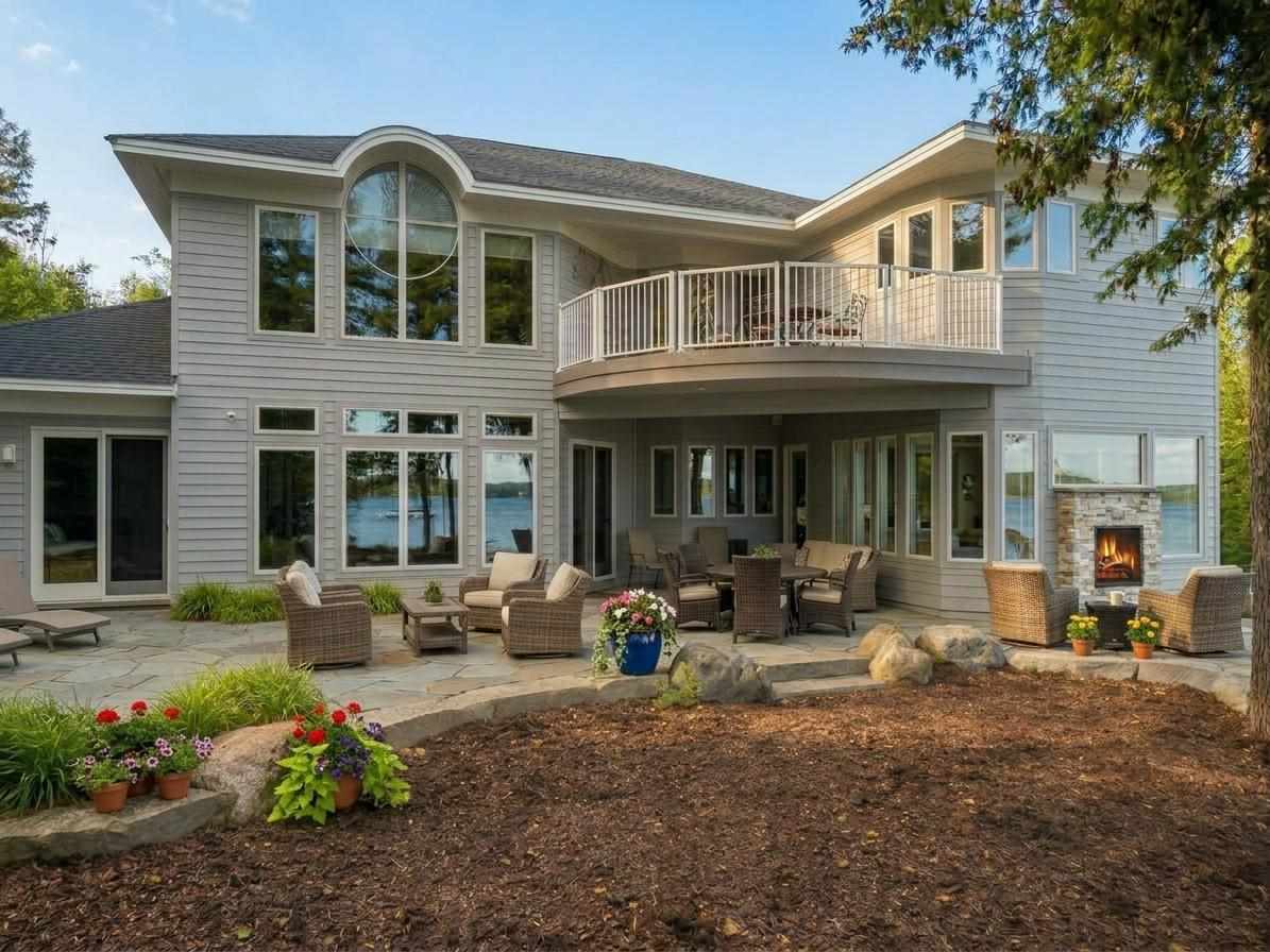 Gray lakeside home with a raised balcony, furnished patio, and outdoor fireplace.
