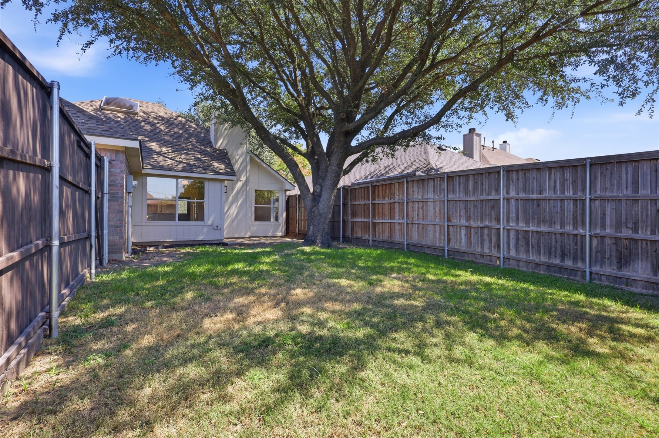 Fenced backyard with a broad lawn, large shade tree, and the rear of a house.